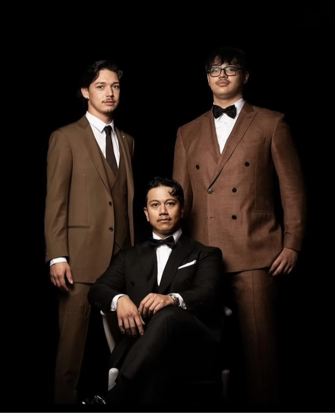 Three men dressed in formal suits against a black background, one seated and two standing, all looking at the camera.