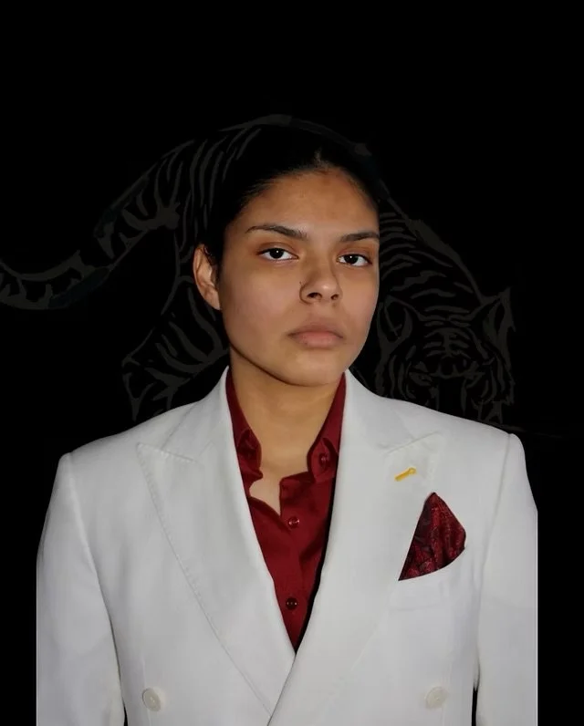 A woman wearing a white blazer over a red shirt with a pocket square, standing against a black background with a tiger graphic.