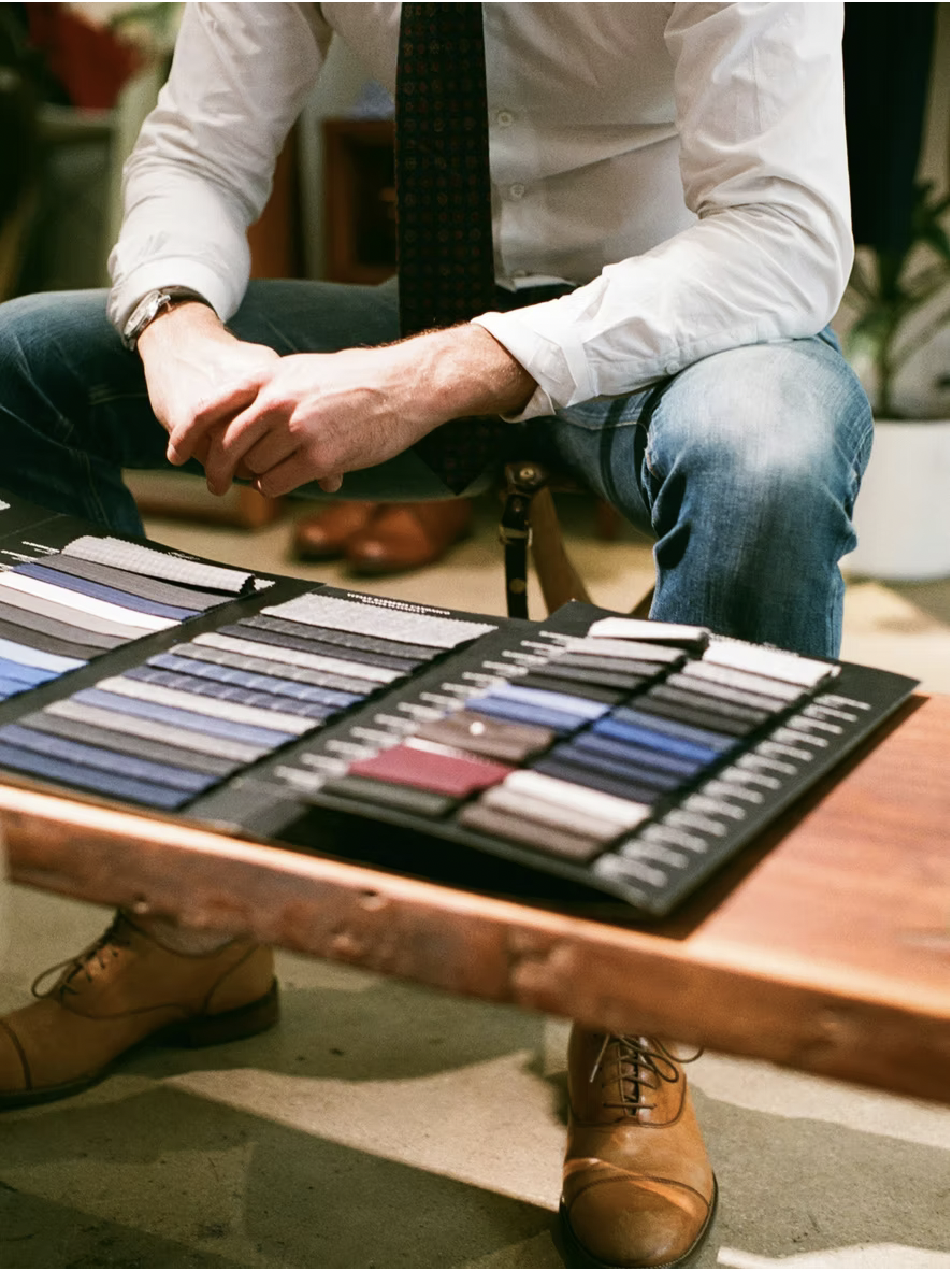 Man wearing white shirt, patterned tie, blue jeans, and brown shoes sitting at a table with fabric swatches.