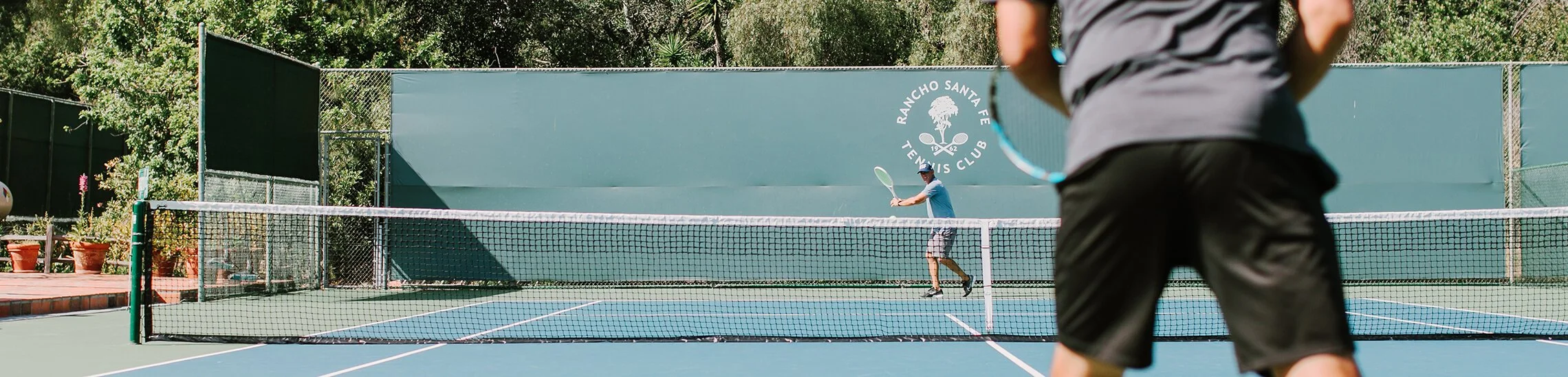 People playing tennis on a court at Rancho Santa Fe Tennis Club, with trees and potted plants nearby.