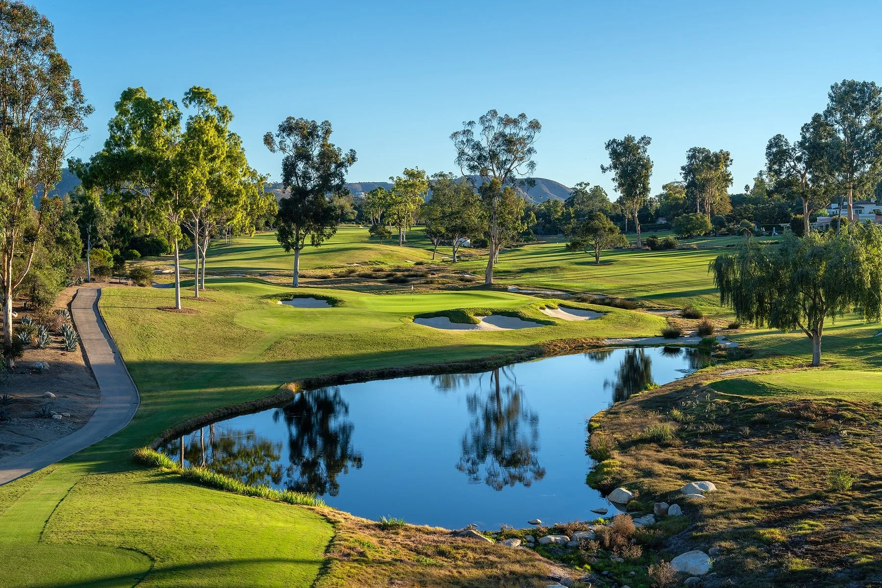 Scenic view of a golf course with lush green grass, sand bunkers, a water hazard, tall trees, and distant mountains under a clear blue sky.