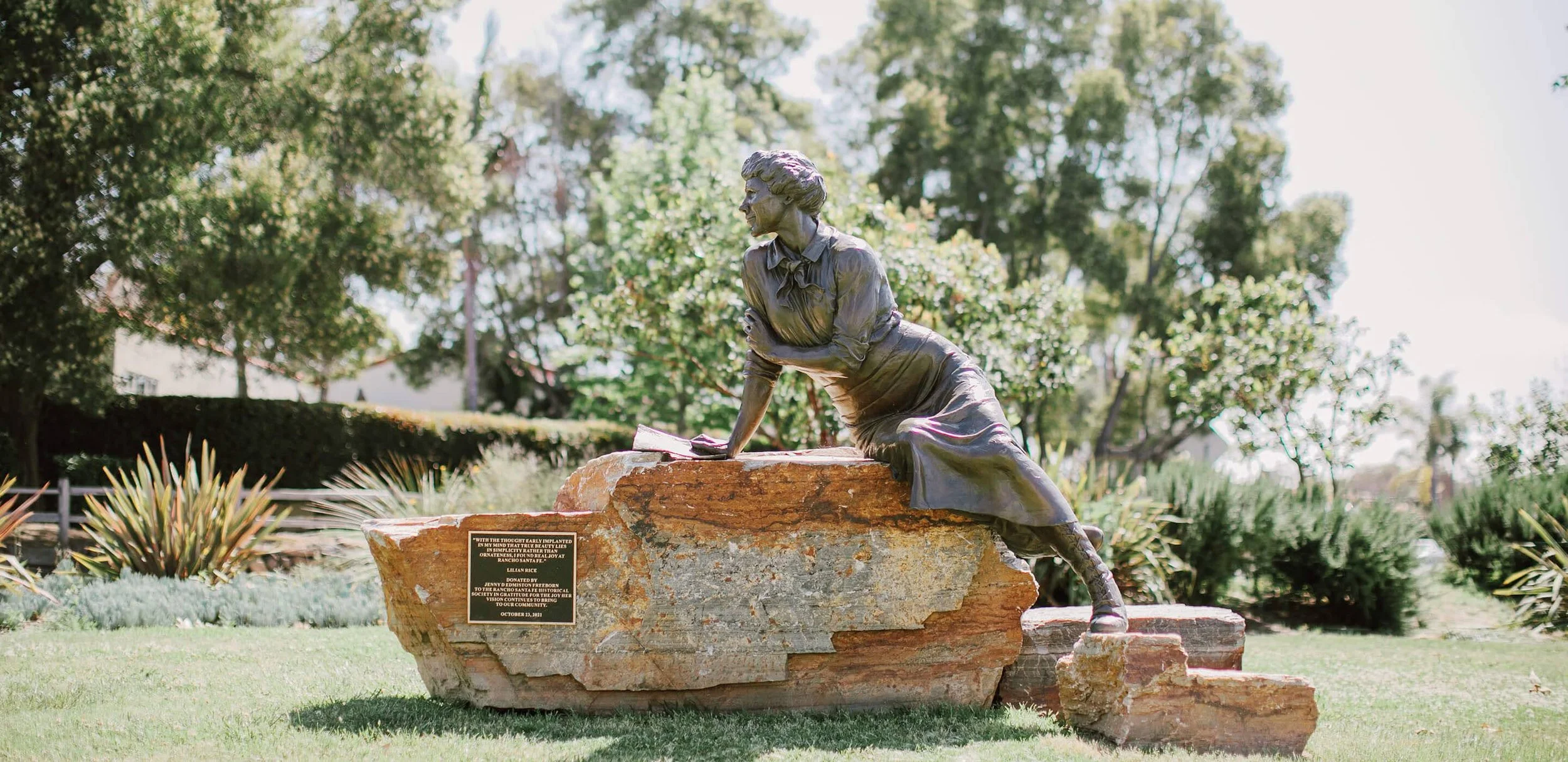 Statue of a woman in a dress kneeling on a rock with a book in her hand, outdoors in a park-like setting with trees and plants.