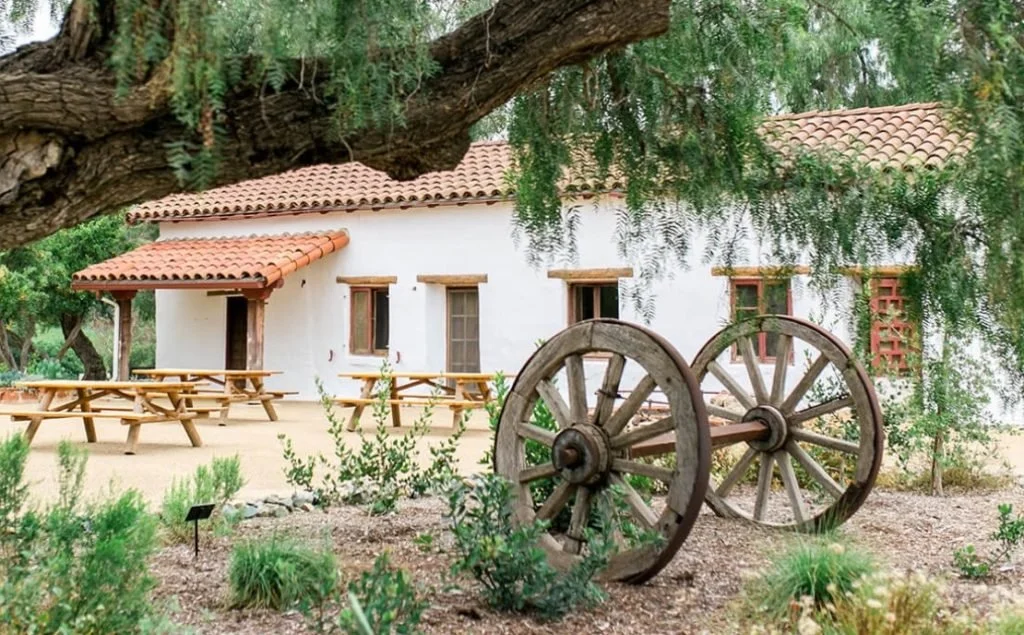A white stucco building with a red tile roof, surrounded by trees and greenery, with outdoor wooden picnic tables and an old wooden wagon wheel in the foreground.