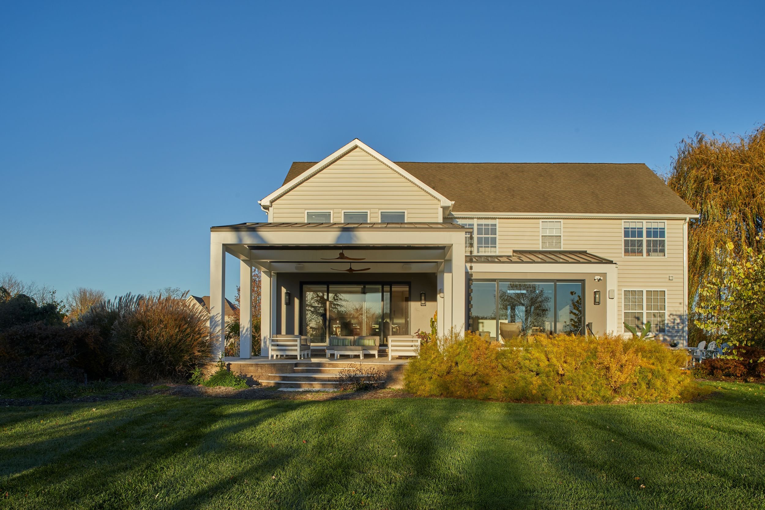 Straight-on rear elevation of a renovated 1990s developer home in Mullica Hill NJ, showing the new covered lanai and structural grid beside the retained original house, overlooking a deep lawn