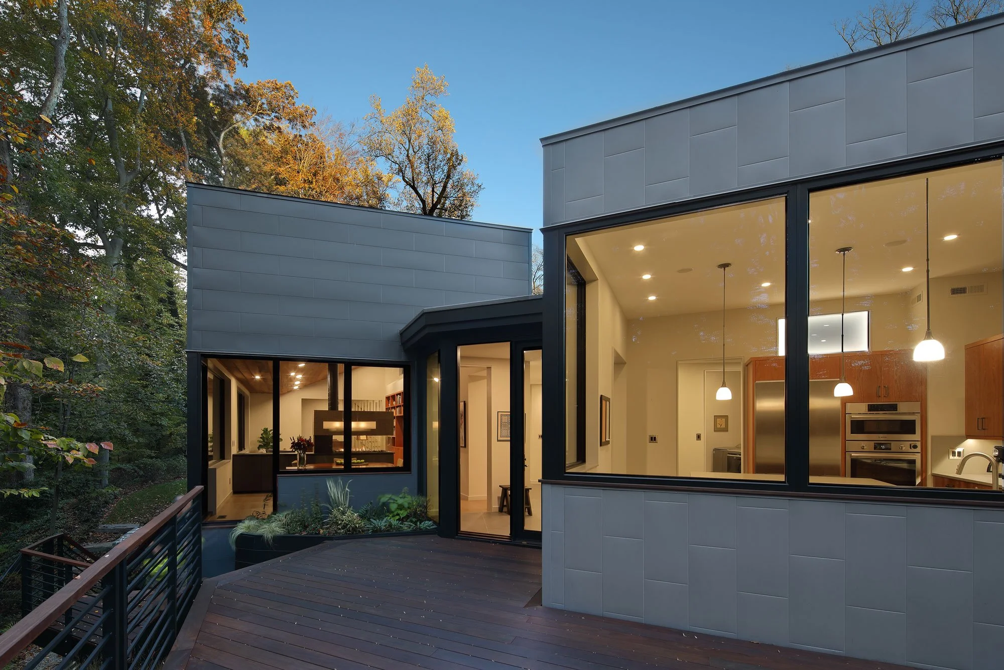 Rear deck of Hadro House Haddonfield NJ at dusk — glowing kitchen volume, ipe decking above the stream bank, deck sized for reading and grilling with family