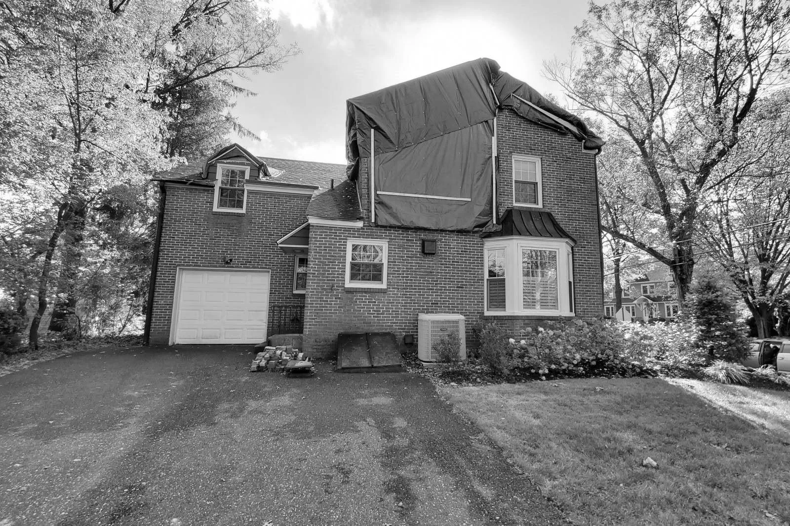 Before photo of a storm-damaged brick center hall colonial home in Haddonfield, NJ showing the side driveway, attached garage, roof tarp, and exterior conditions before renovation.