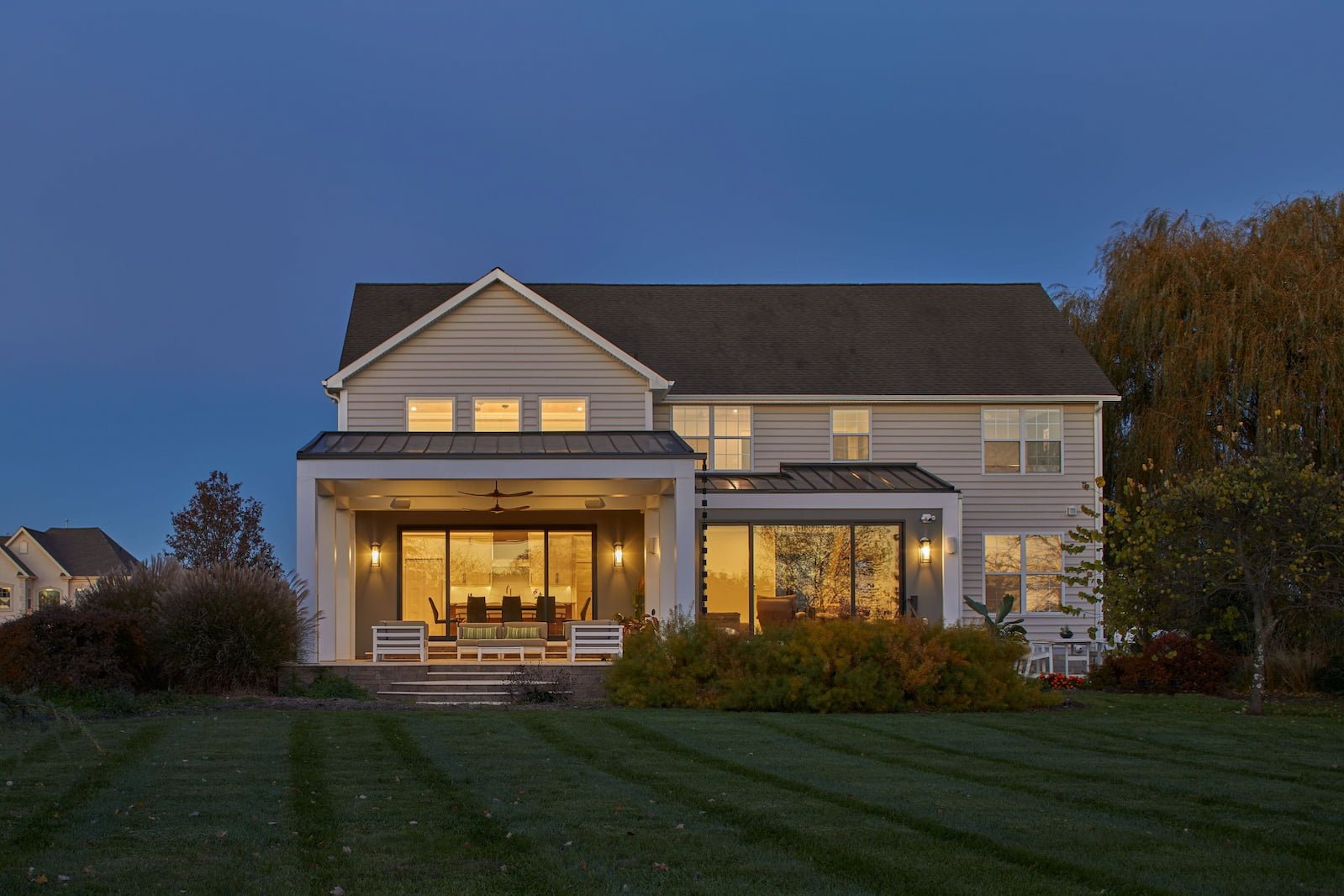 McMansion Makeover rear addition at dusk Mullica Hill NJ — 1990s developer home renovation covered porch floor-to-ceiling glass standing seam metal roof indoor-outdoor living yard connection J Reinert Architecture