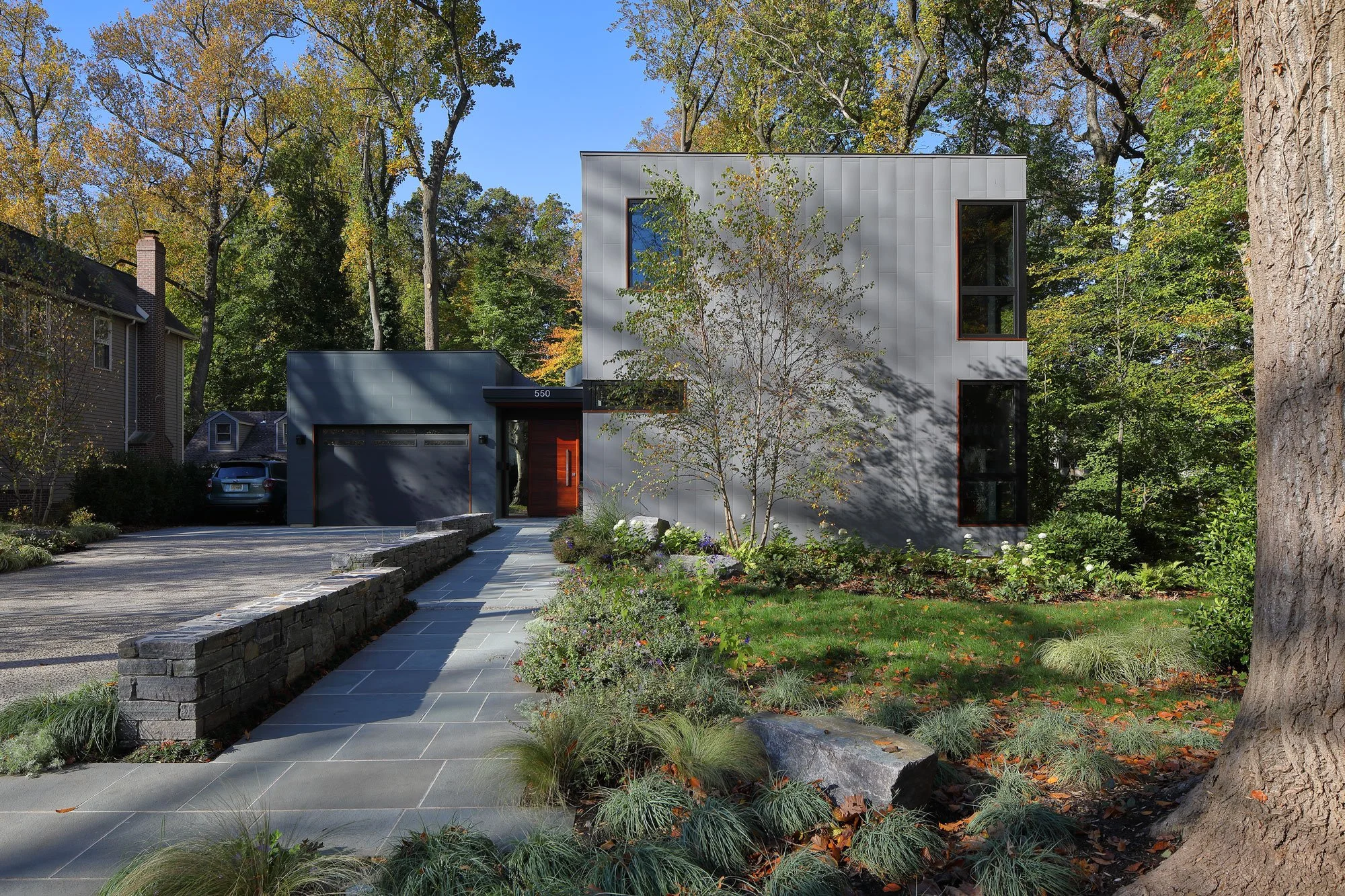 Arrival court at Hadro House Haddonfield NJ — inclined bluestone walk, stone site walls, and native plantings leading to the wood pivot entry door, step-free by design