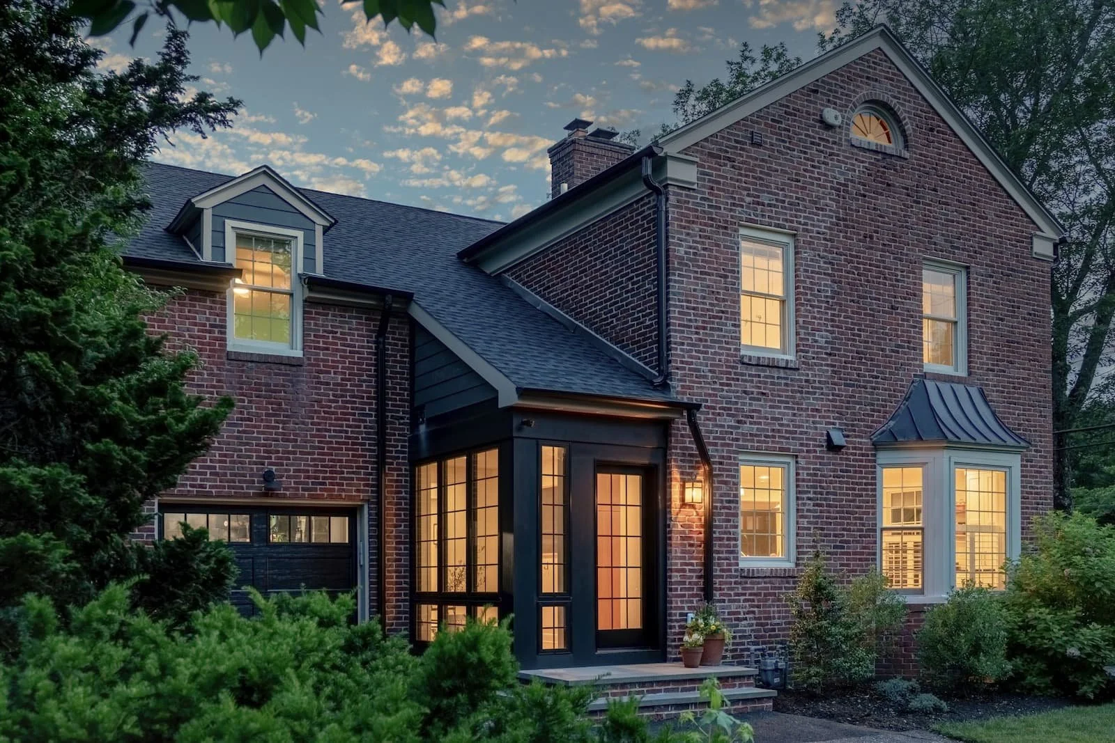 Center Hall Industrial Minimalism renovation Haddonfield NJ — 1930s brick colonial dusk contemporary black fiberglass grid entry pavilion standing seam metal bay window warm interior light landscaping J Reinert Architecture