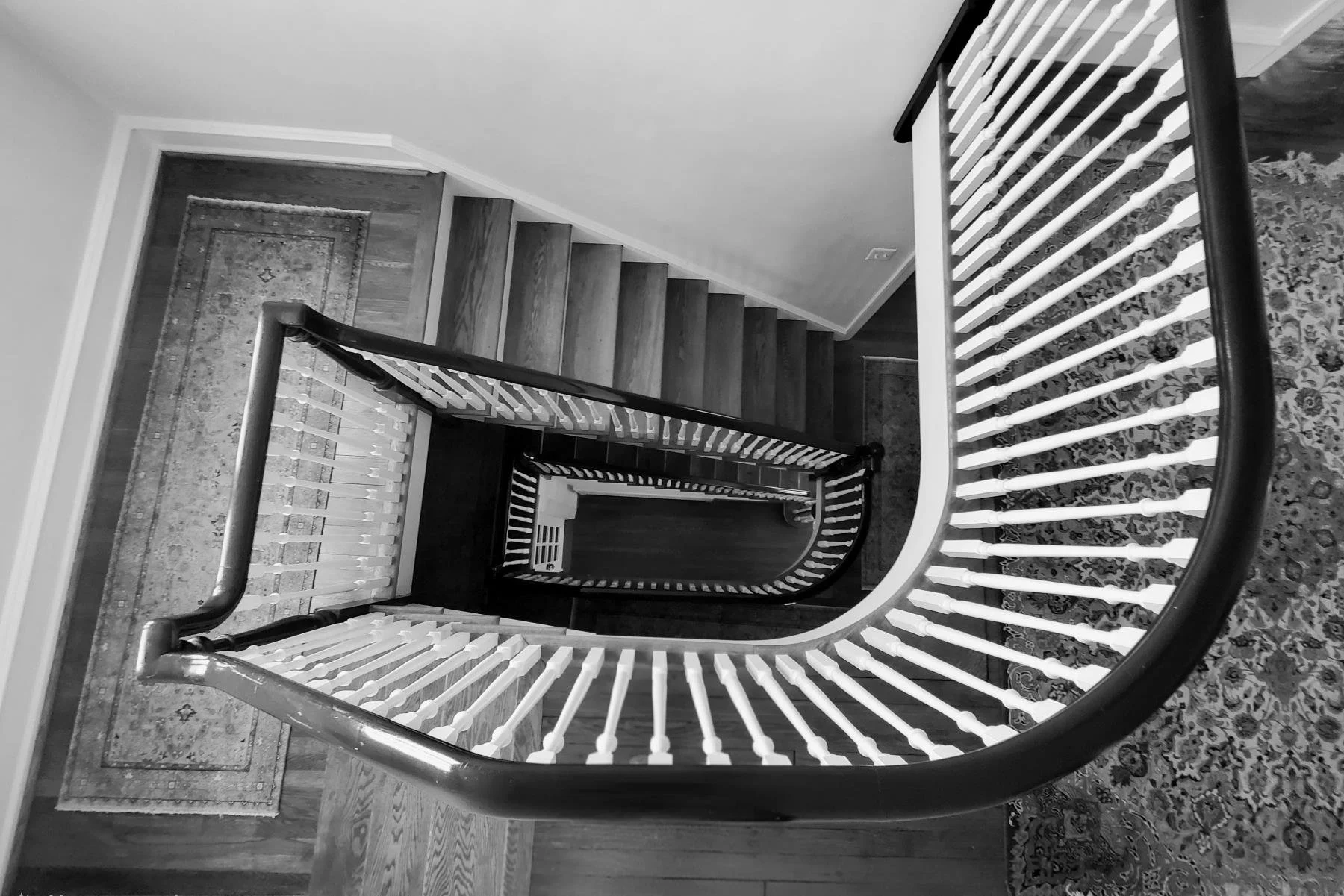 Black-and-white view down a historic home’s staircase, showing original wood treads, curved handrail, white balusters, patterned rugs, and traditional architectural detailing.