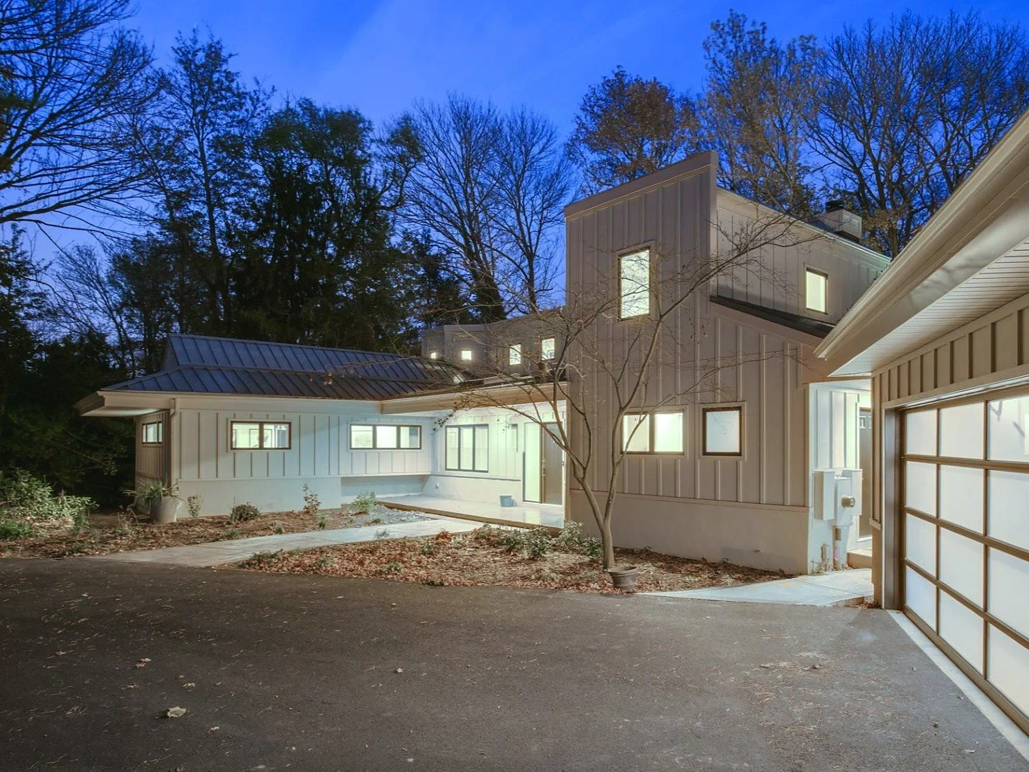 Twilight view of a renovated mid-century modern home in Cherry Hill, NJ, with a board-and-batten upper volume, standing-seam metal roof, and a backlit translucent garage door.