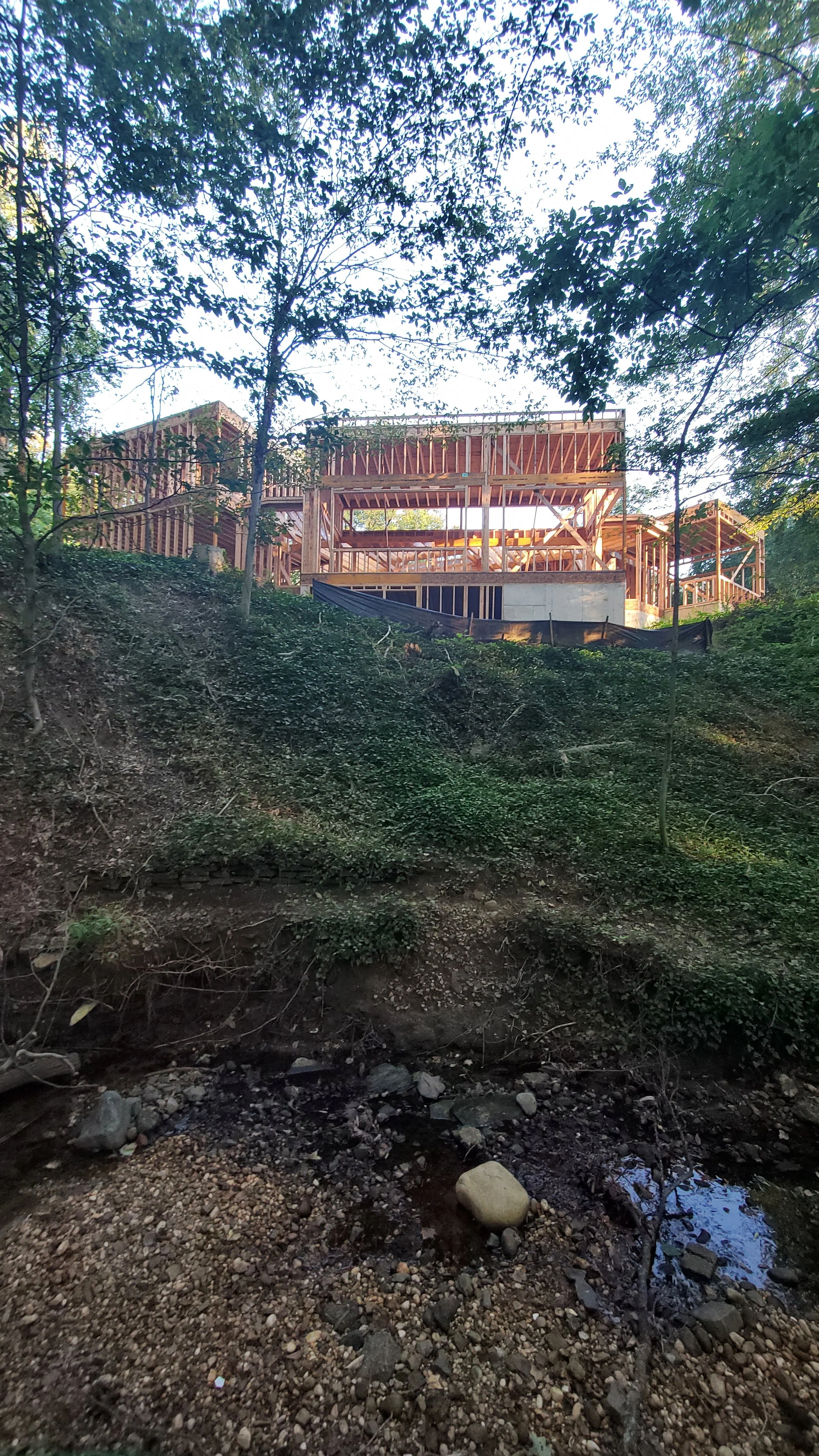 Hadro House framing seen from the stream bed below in Haddonfield NJ — volumes rising above the protected ravine slope with erosion controls in place during construction