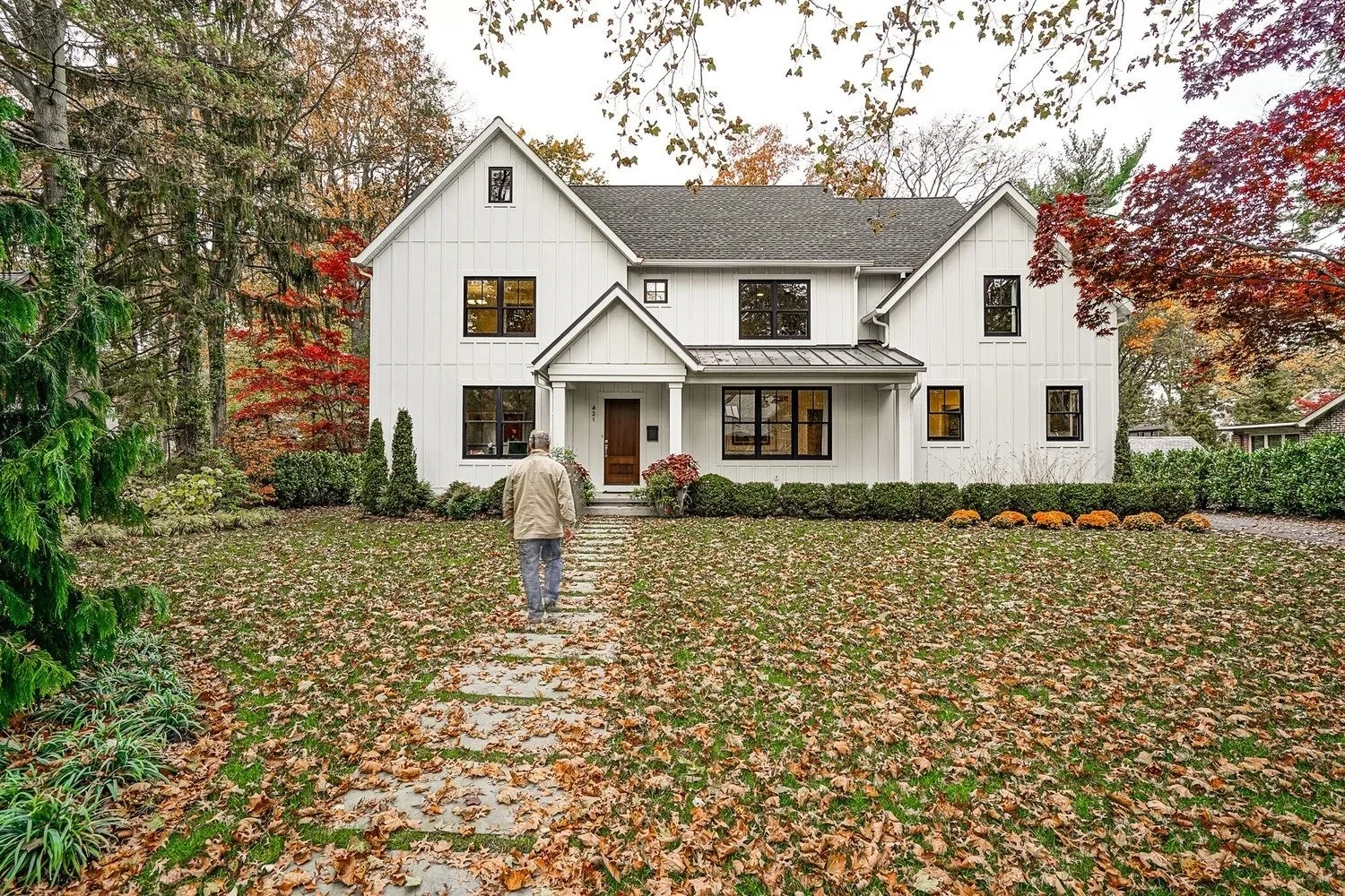 Person walking along a stone path toward a white custom home with black windows, autumn leaves, and layered landscaping.