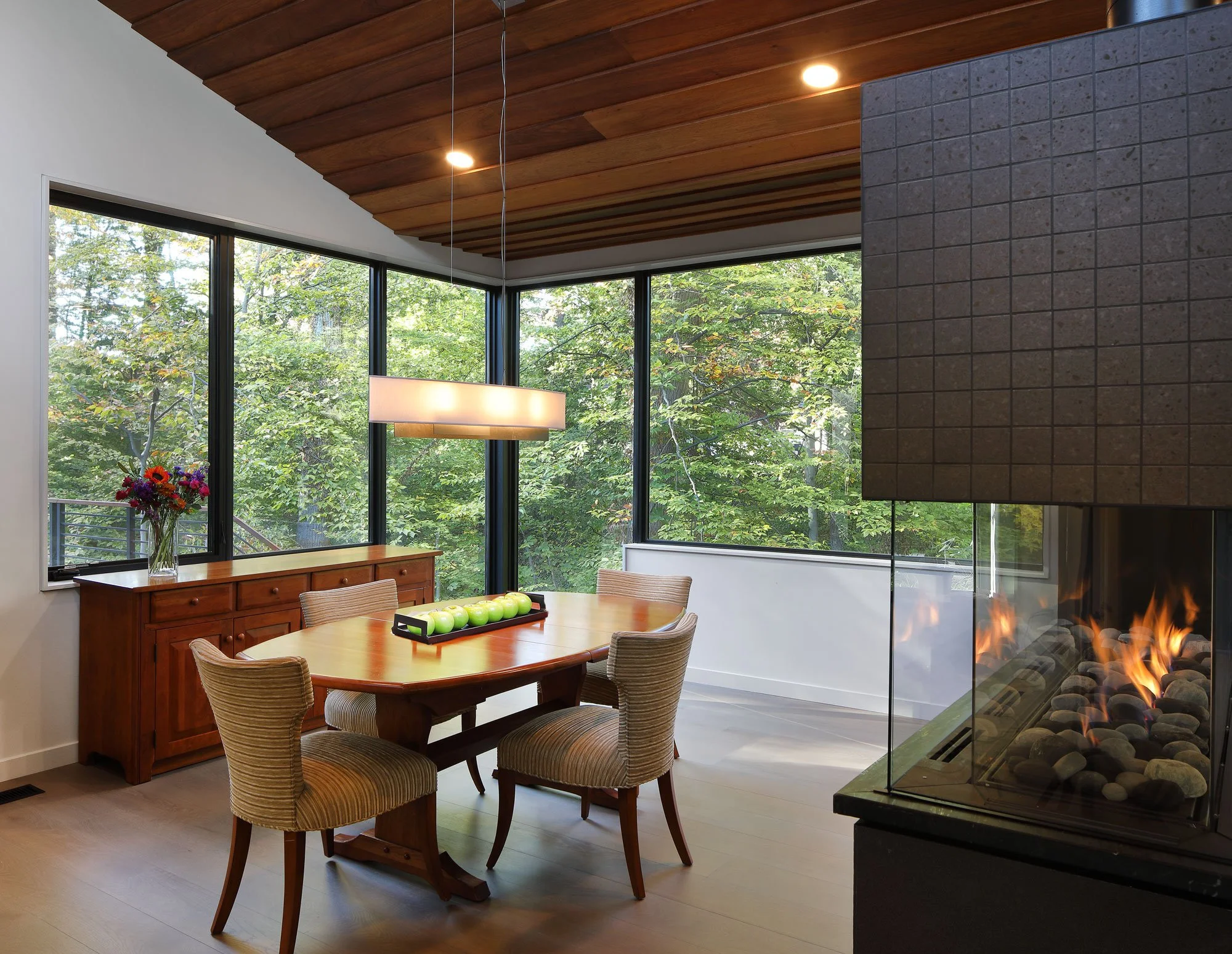 Three-sided gas fireplace at Hadro House in Haddonfield NJ dividing dining and living spaces without closing them, with corner glass walls facing the wooded stream bank