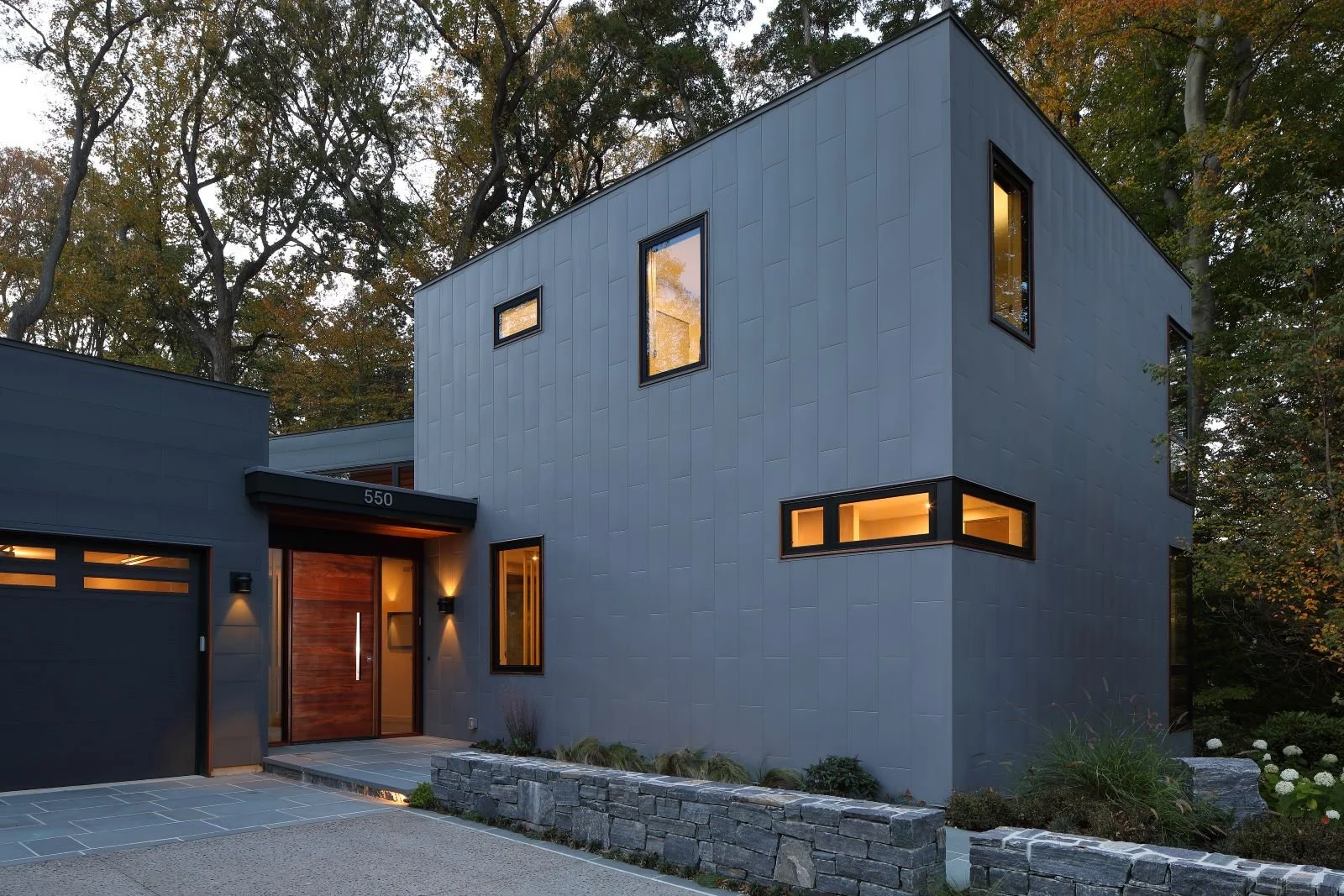 Front entry of Hadro House, a contemporary gray-clad home in Haddonfield, New Jersey, with black-framed windows, a warm wood door, and integrated landscape walls at dusk.