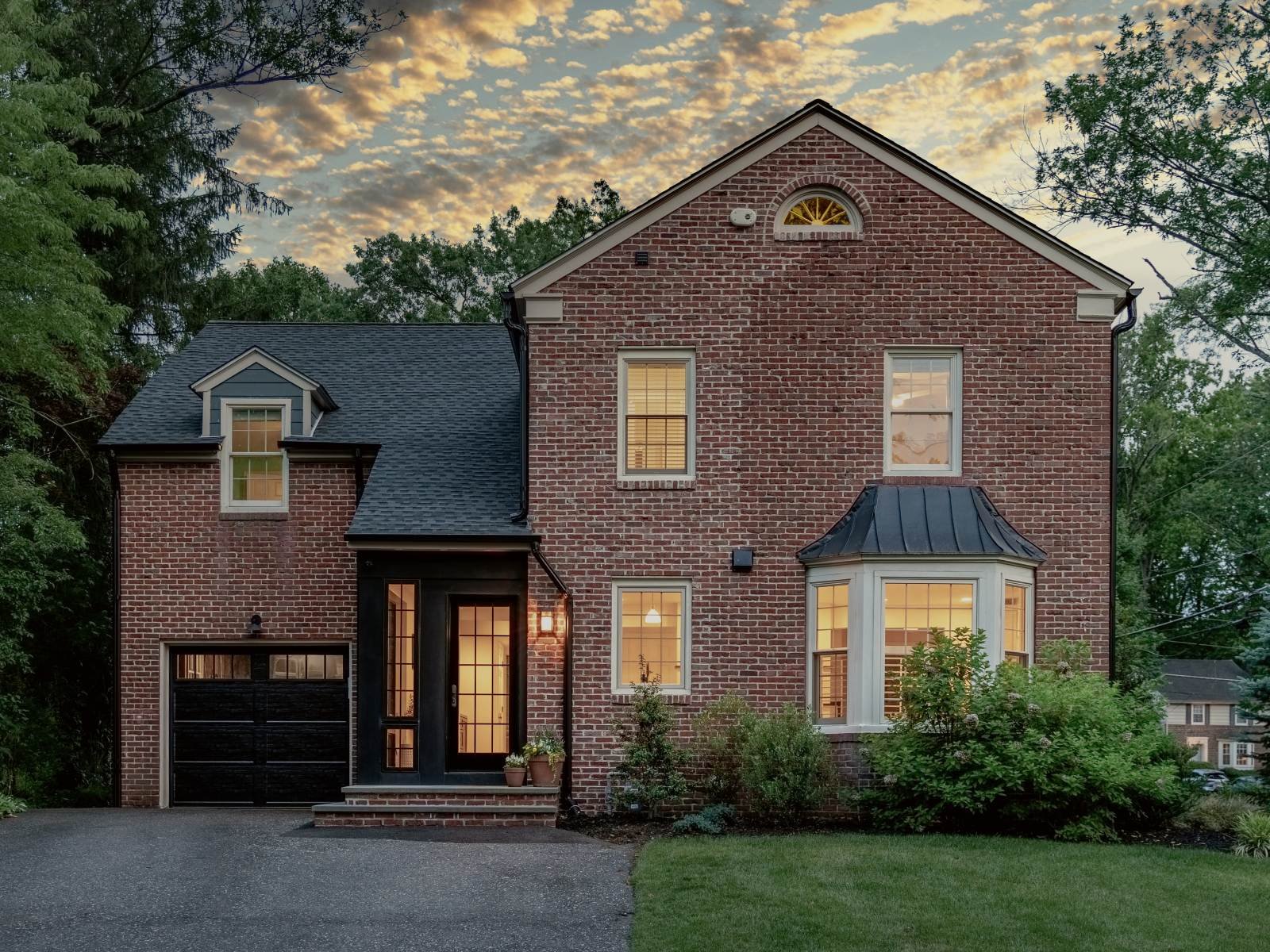 Dusk front elevation of a renovated 1930s brick center-hall colonial with a standing-seam bay roof, black entry surround, and warm interior light, Haddonfield, NJ.