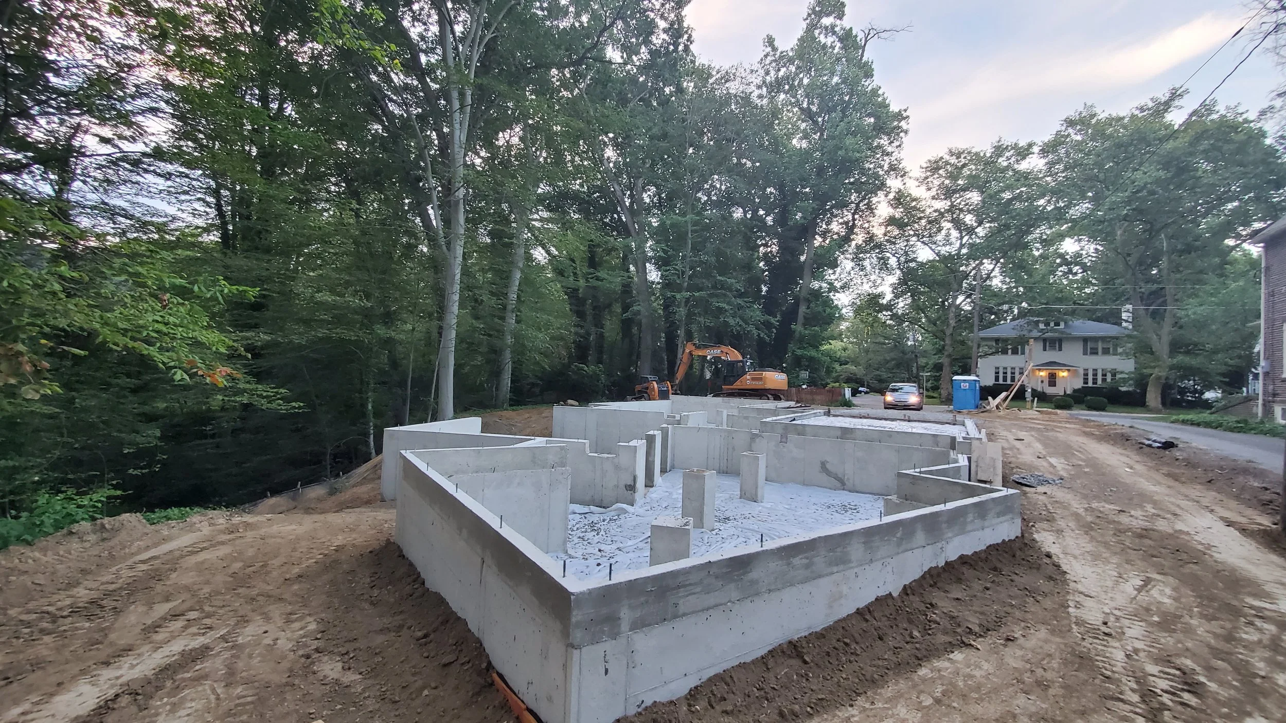 Concrete foundation walls of Hadro House in Haddonfield NJ at dusk, July 2020 — the plan of offset volumes legible in fresh concrete beside traditional neighboring homes