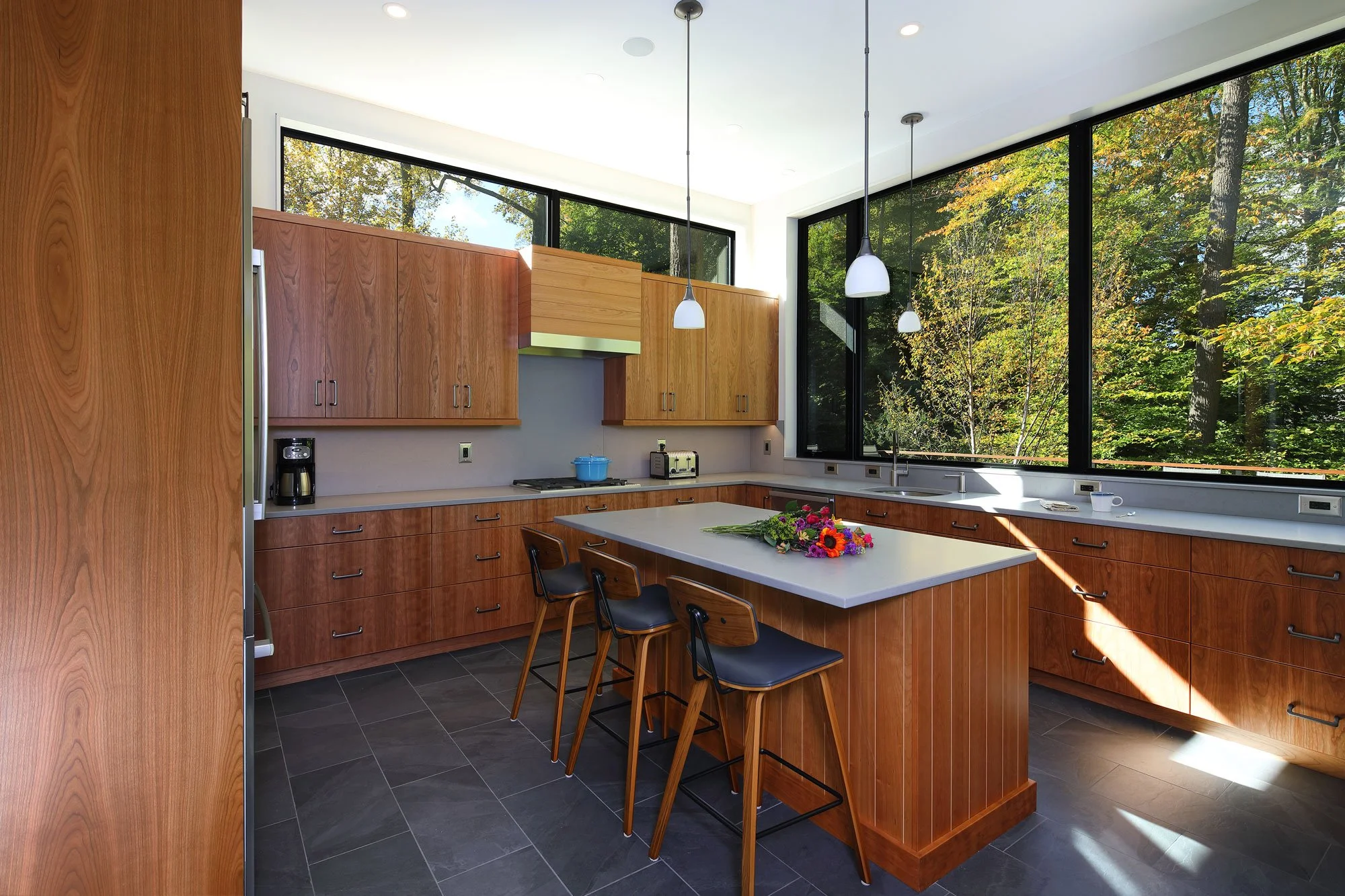 Kitchen at Hadro House Haddonfield NJ — warm wood slab cabinets, island with counter seating for three, and continuous windows to the treetops