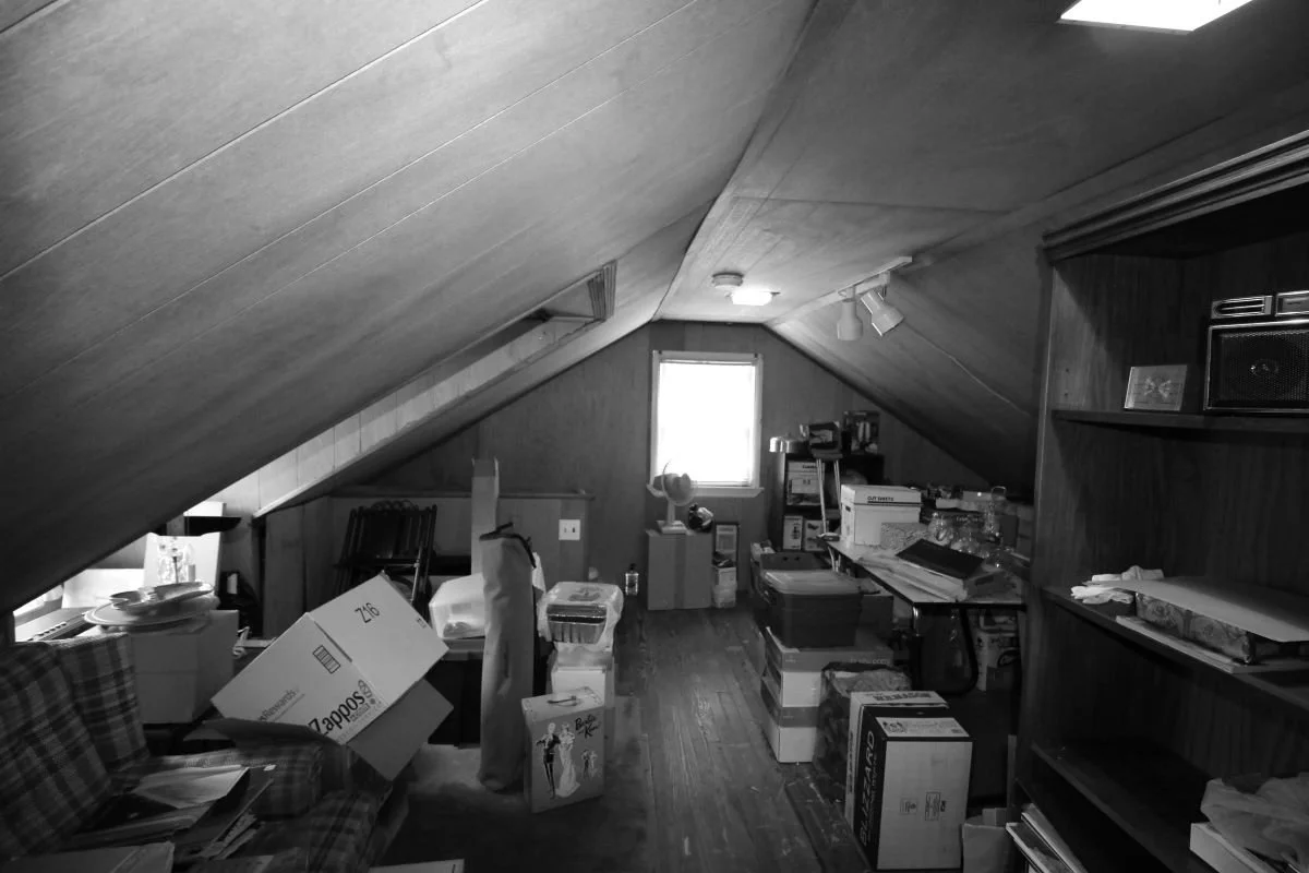 Before renovation — unfinished attic storage at cape cod home in Haddon Heights NJ showing low paneled sloped ceiling, boxes and furniture packed throughout, and one small dormer window with minimal light, prior to J Reinert Architecture renovation