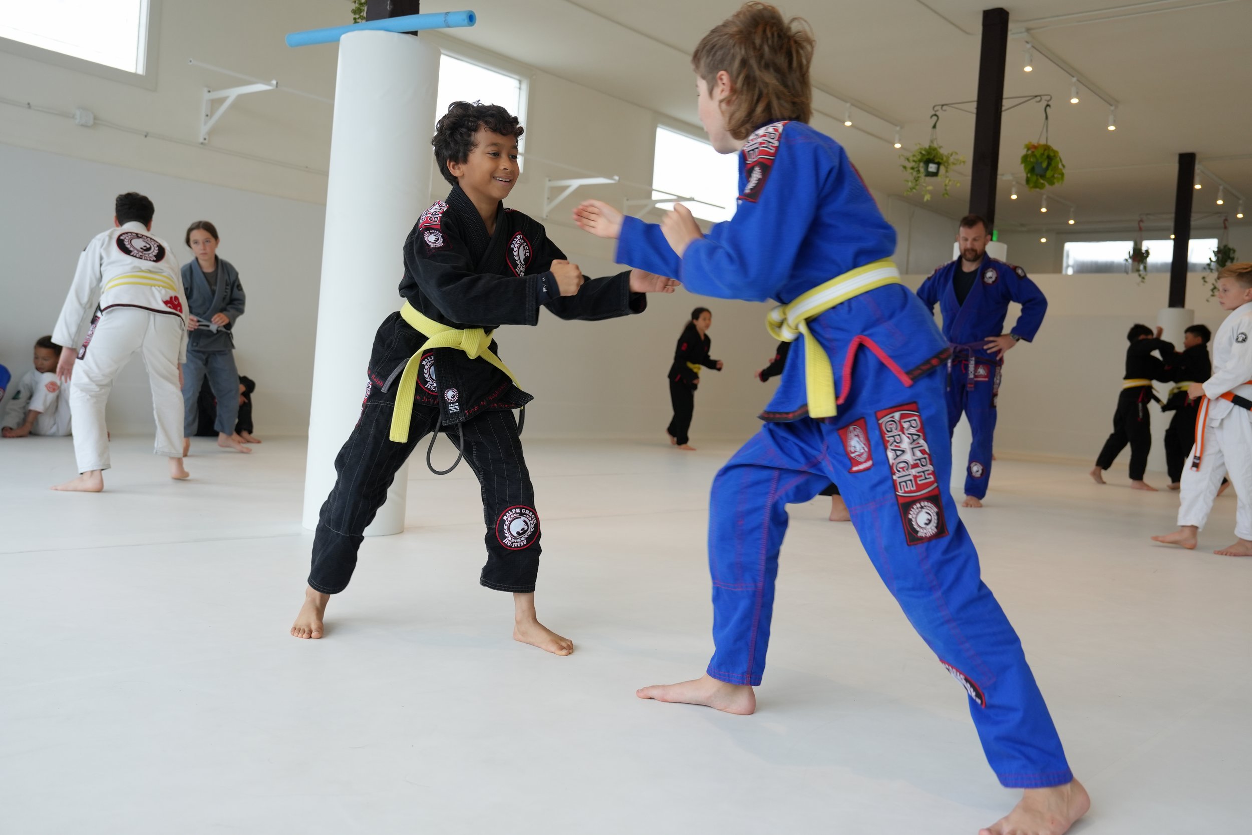 Two children in Brazilian Jiu-Jitsu uniforms practicing a grip fight, or 'pummel' drill, during a BJJ class in a dojo with other students and instructors in the background.