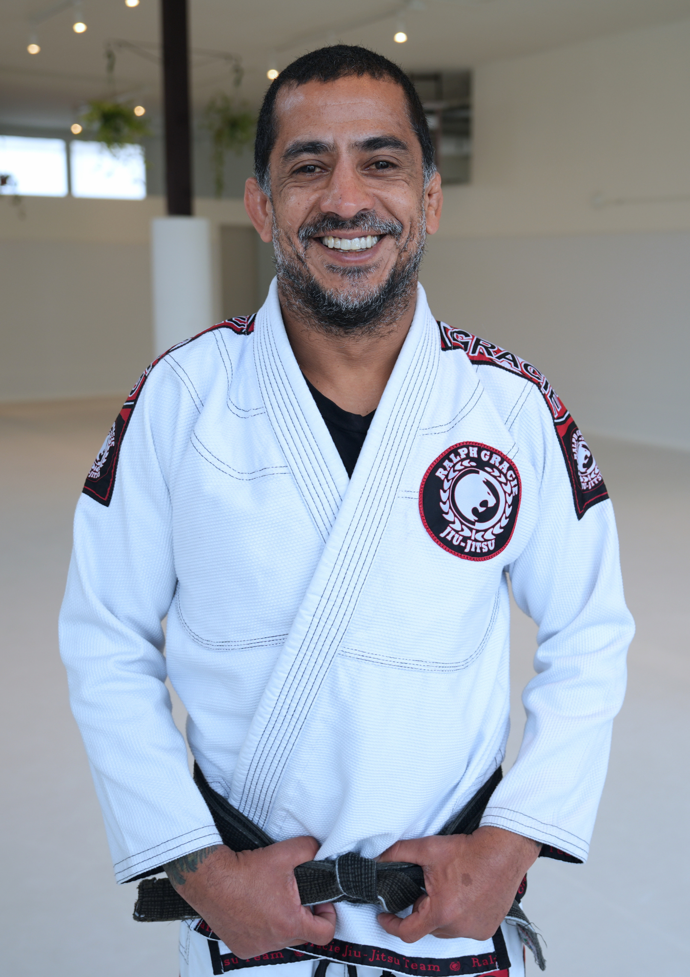 A smiling man wearing a white Brazilian Jiu-Jitsu gi with patches, standing in a well-lit indoor space.