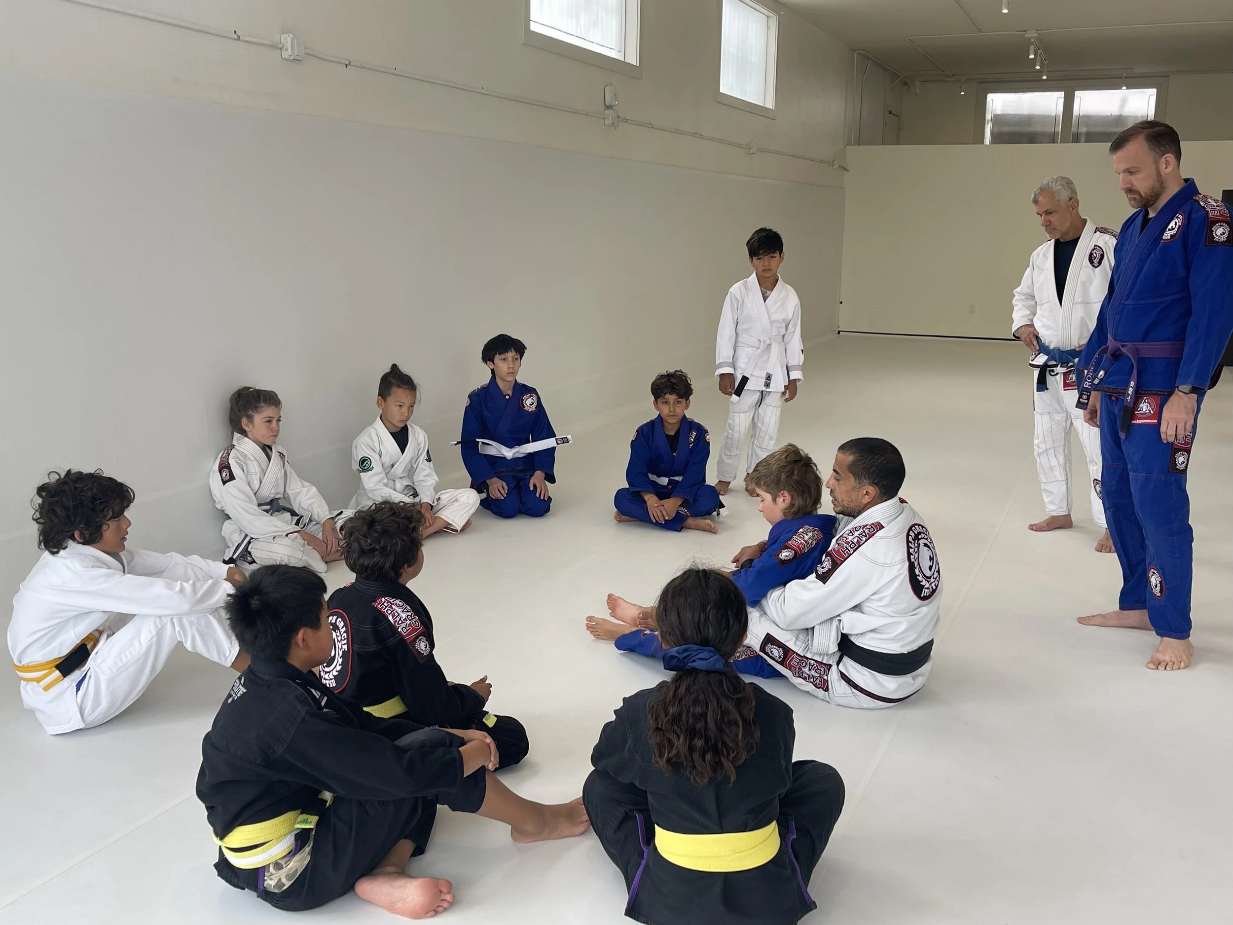Children and instructors sitting on the floor in a martial arts class, wearing gi uniforms with belts, inside a bright training room.