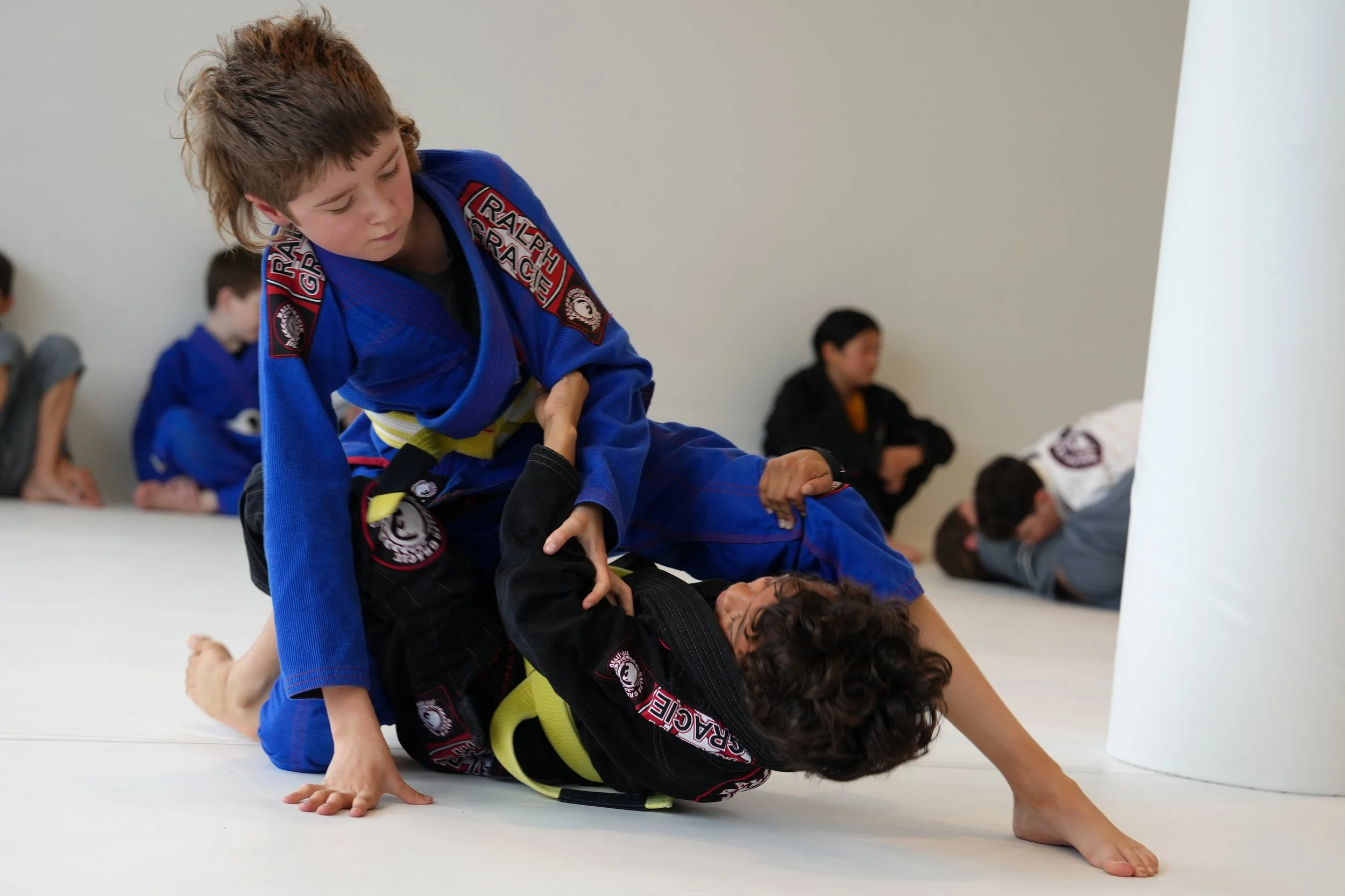 Children practicing Brazilian Jiu-Jitsu on a white mat, with one girl in a blue gi applying a ground technique and others watching in the background.
