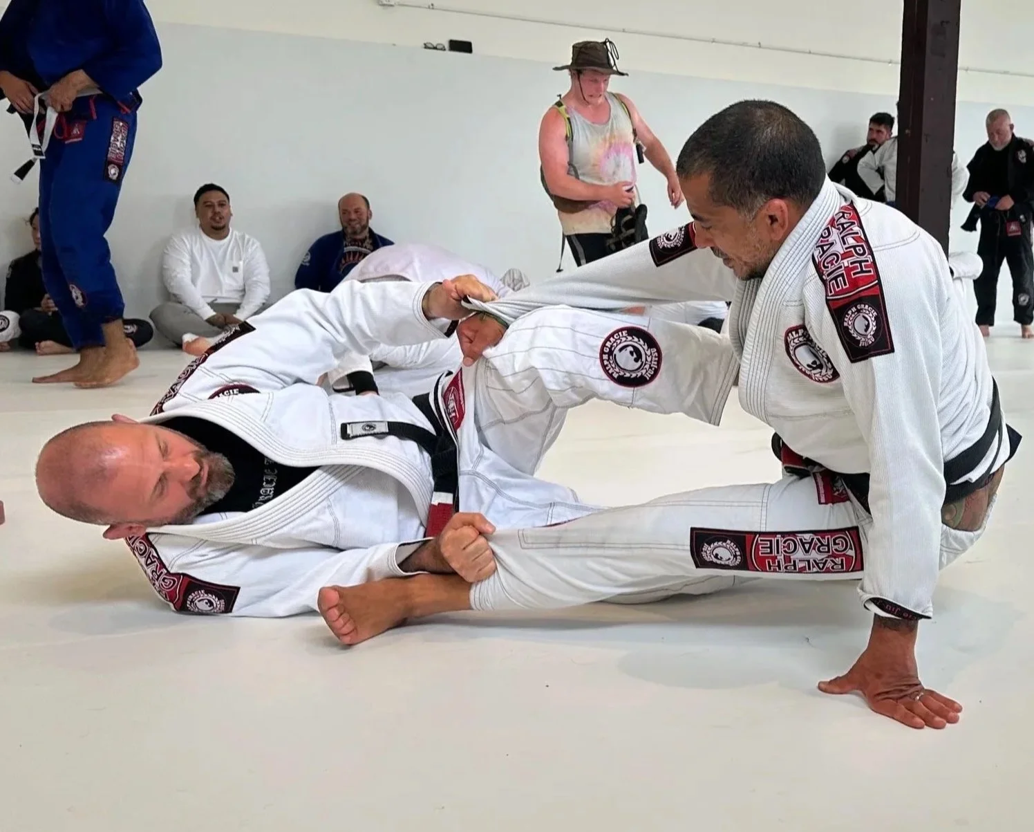 Two men wearing Brazilian Jiu-Jitsu gis are engaged in a grappling match on the mat while others watch in the background, inside a dojo.