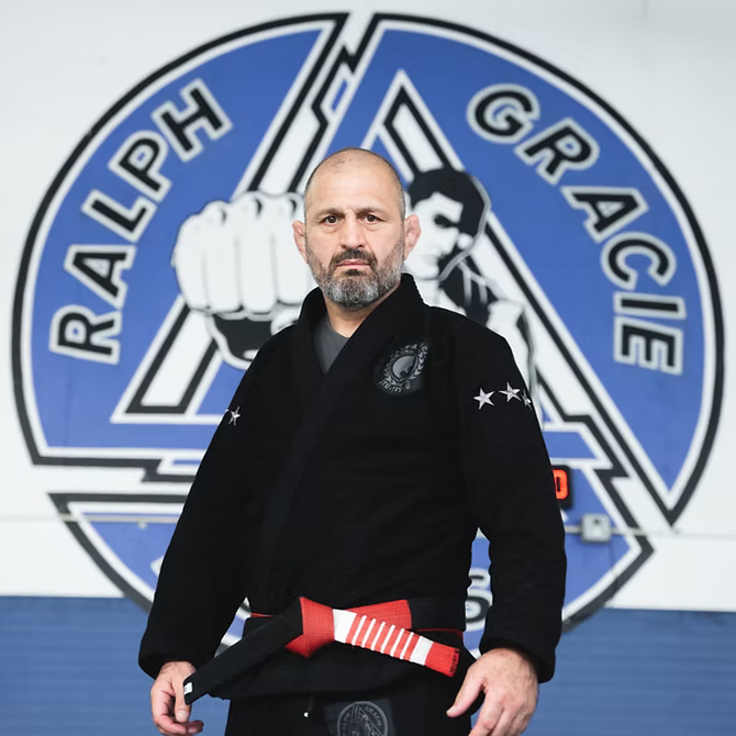 A man in a black Brazilian Jiu-Jitsu gi holding a red and white striped belt, standing in front of a martial arts training facility with a giant logo that reads "Ralph Gracie" and features a clenched fist.