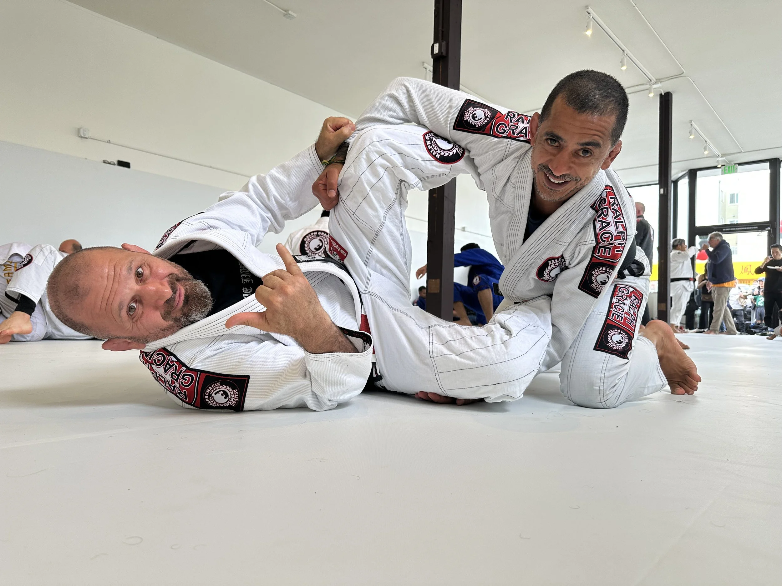 Two men practicing Brazilian Jiu-Jitsu on a white mat; one man in a black Gi, the other in a white Gi, smiling, with an indoor gym setting in the background.