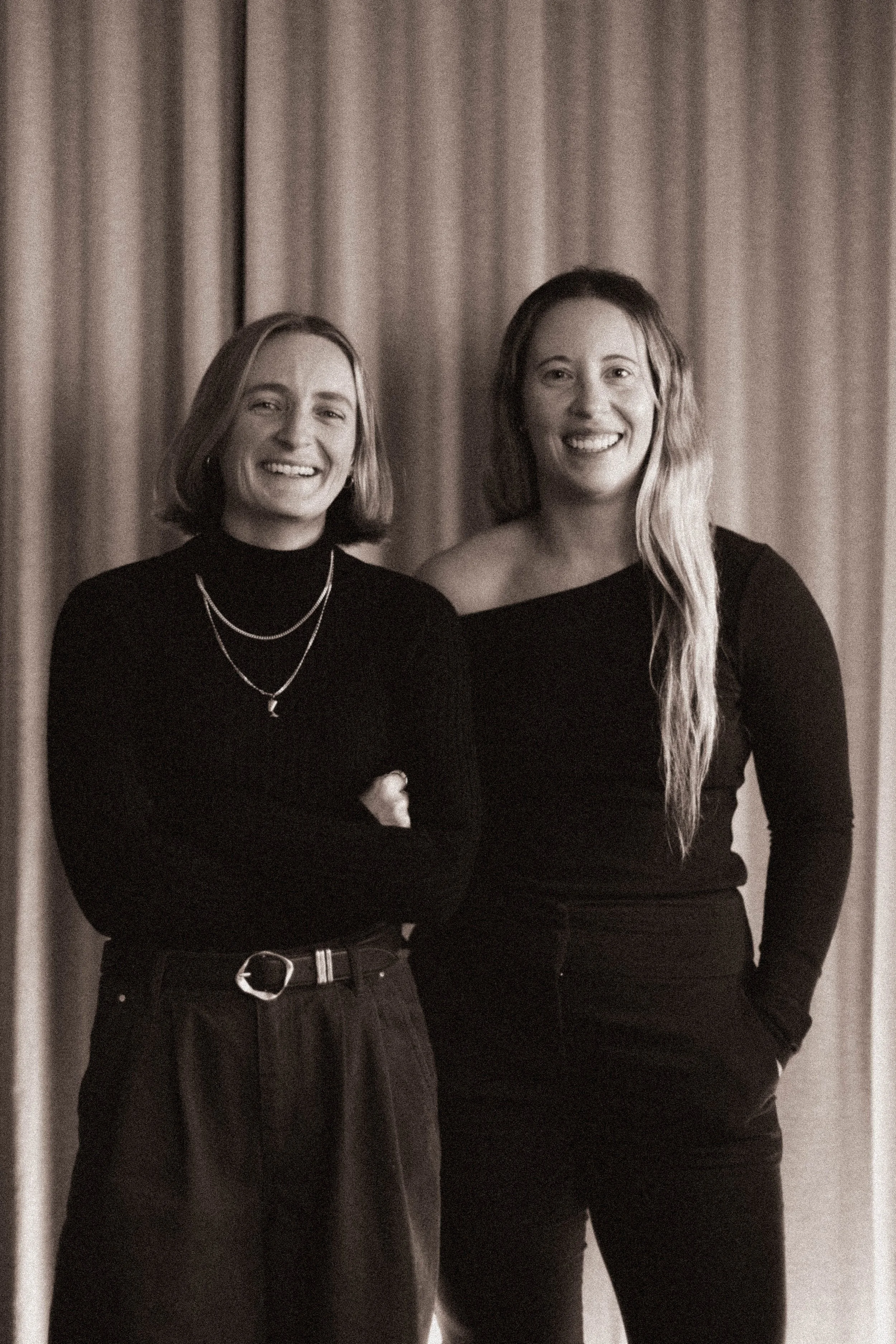 Two women standing side by side, smiling, with a curtain background. They are dressed in dark clothing, one with a turtleneck and layered necklaces, the other with an off-shoulder top. The photo has a black-and-white filter.
