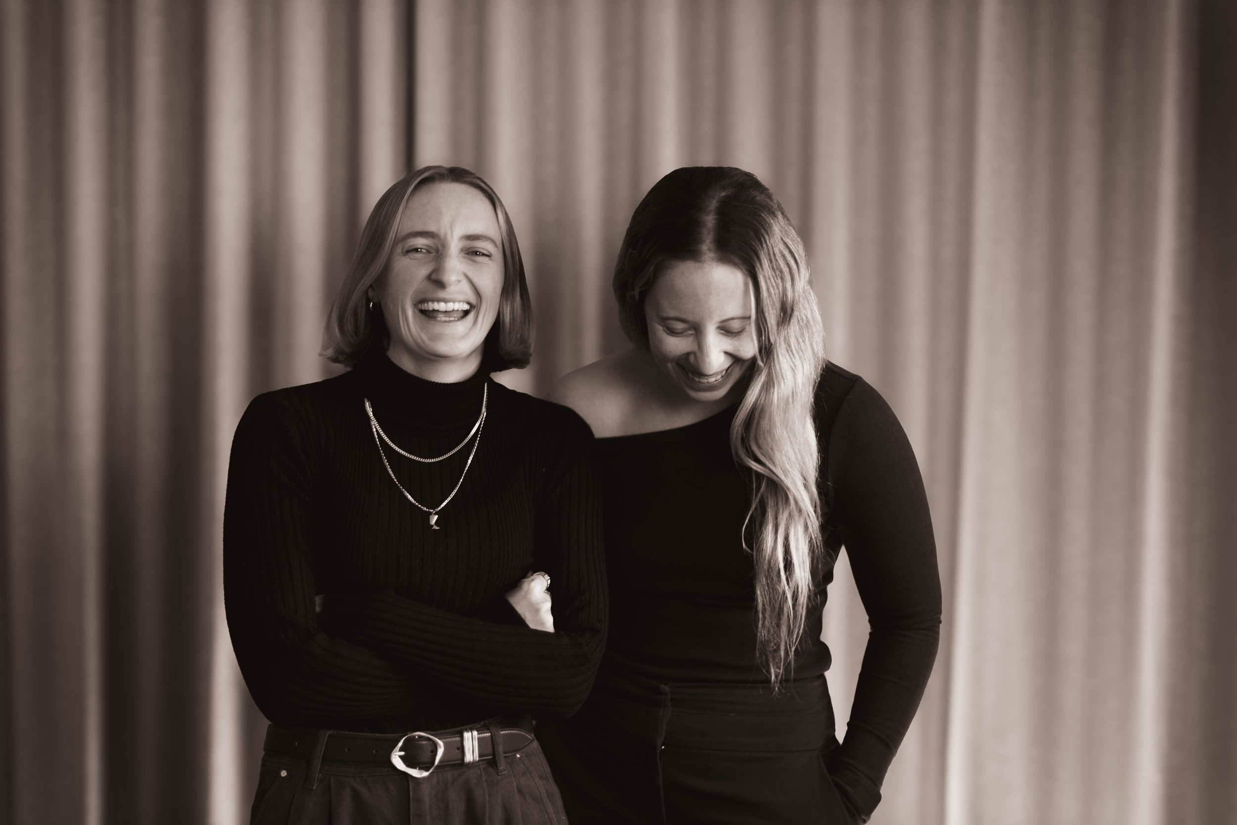 Two women laughing together in front of beige curtains.