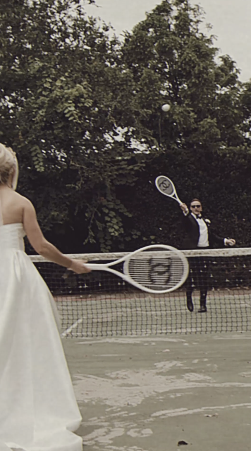 A woman in a white dress is playing tennis on an outdoor court, with a man in a tuxedo and sunglasses in the background hitting a tennis ball with a racket.