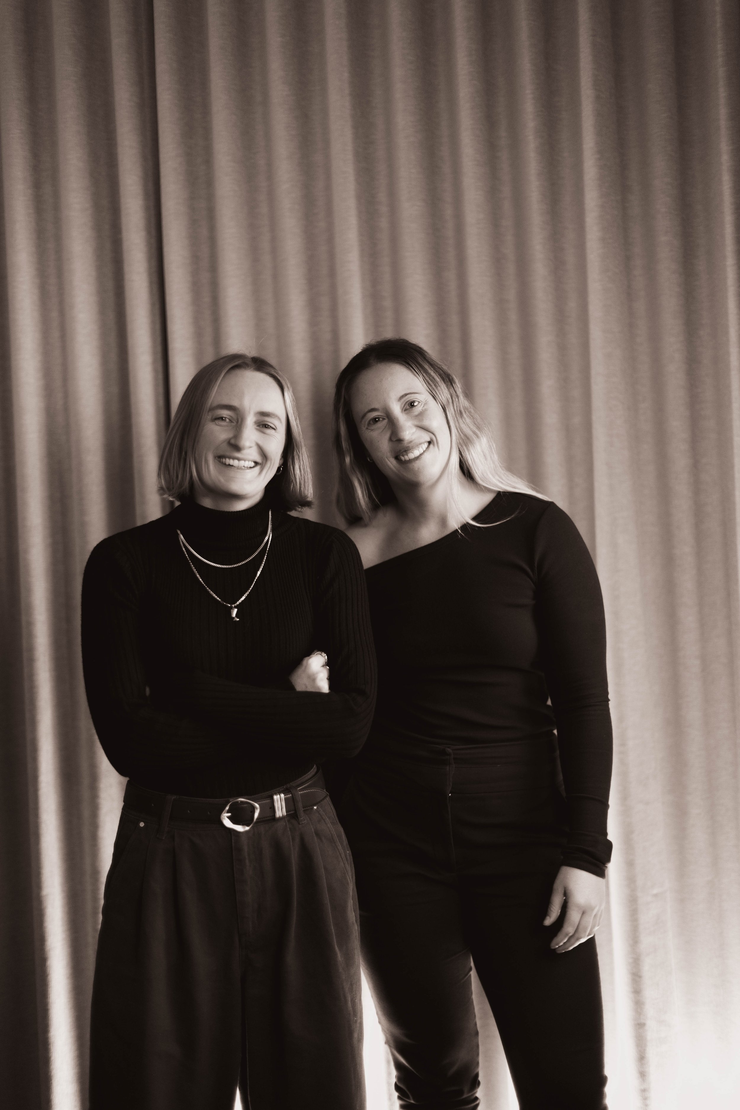 Two women standing in front of a curtain, smiling, dressed in black.