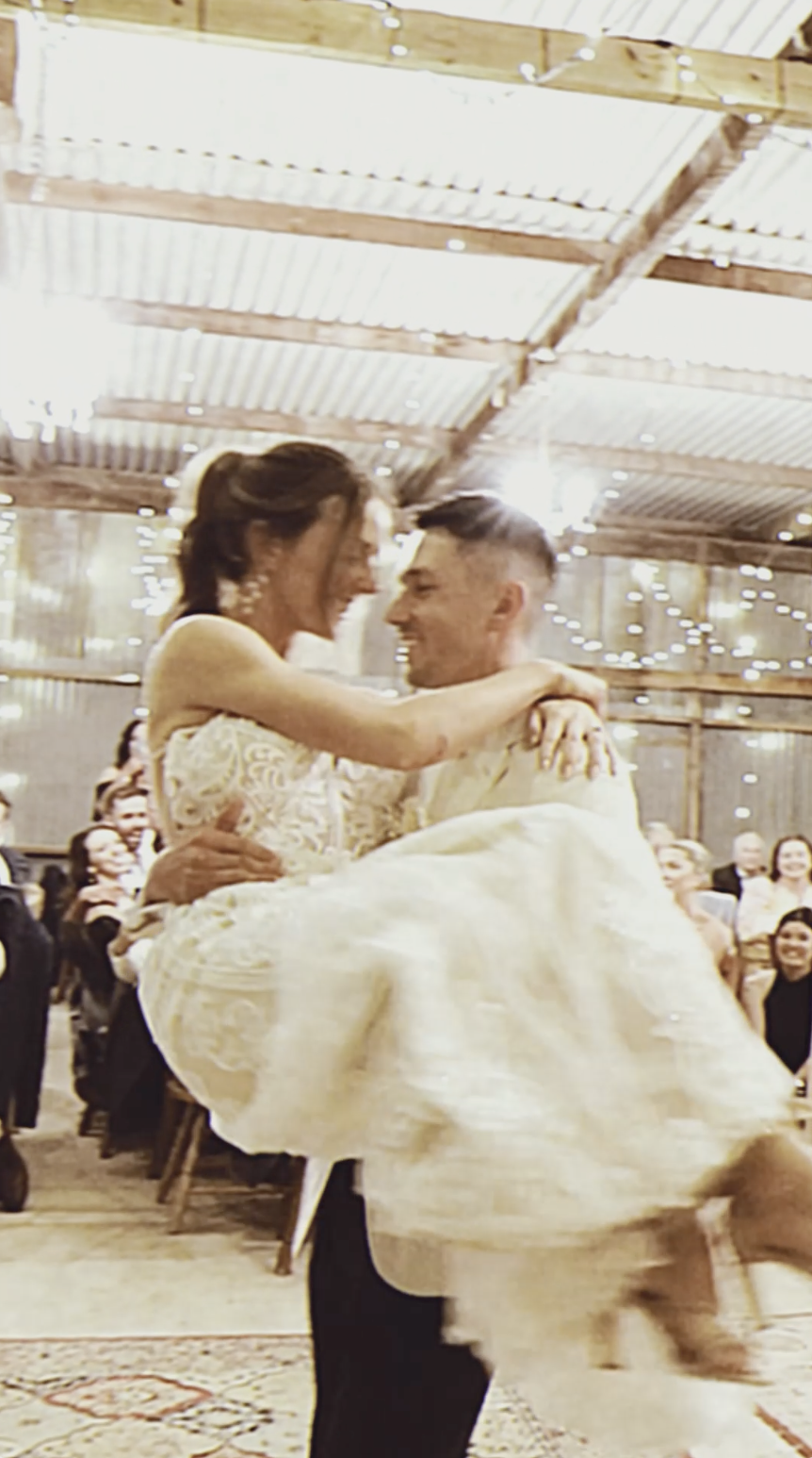 A man carrying a woman in a white lace dress, likely during a wedding dance, inside a rustic venue with string lights and wooden beams.