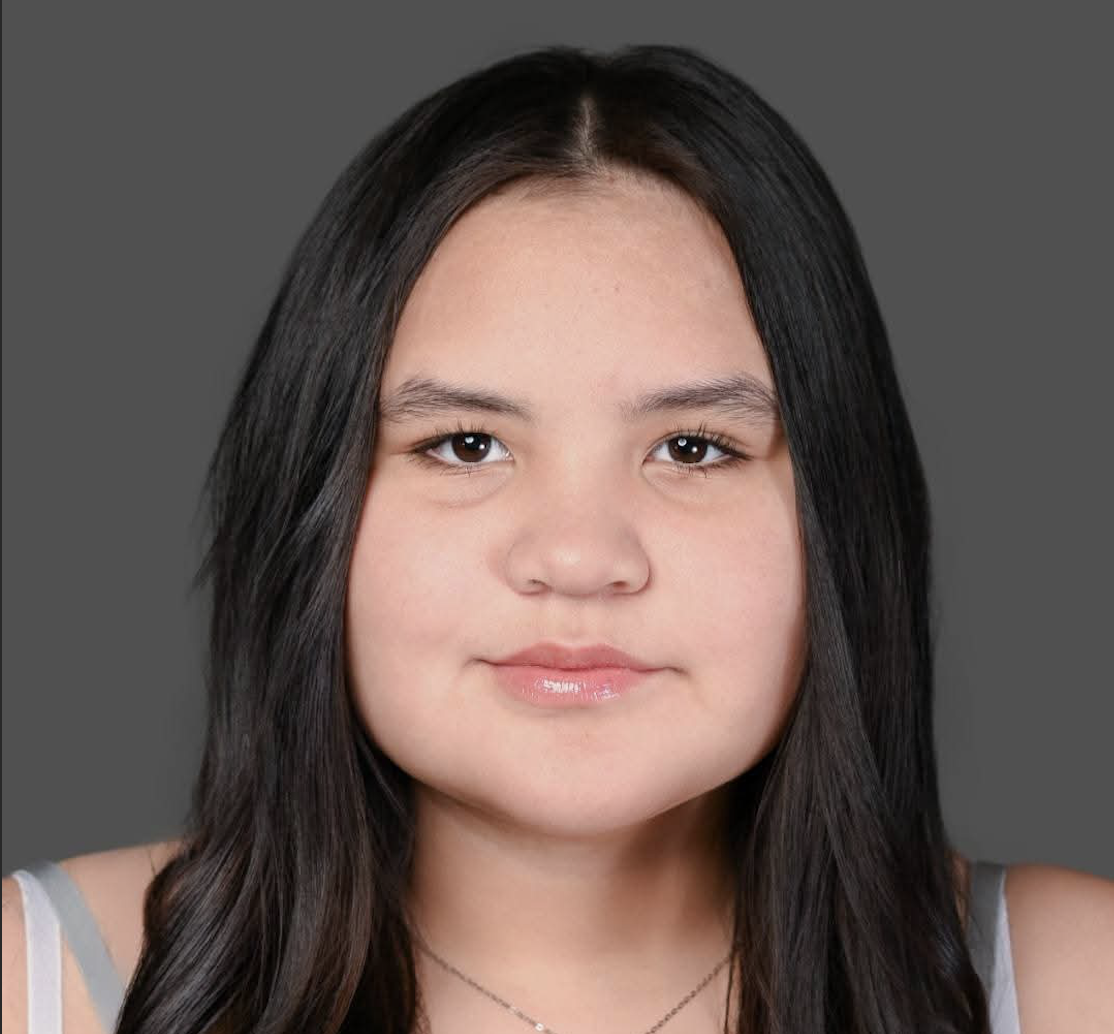 Portrait of a young girl with long dark hair wearing a white top with gray straps, on a gray background.