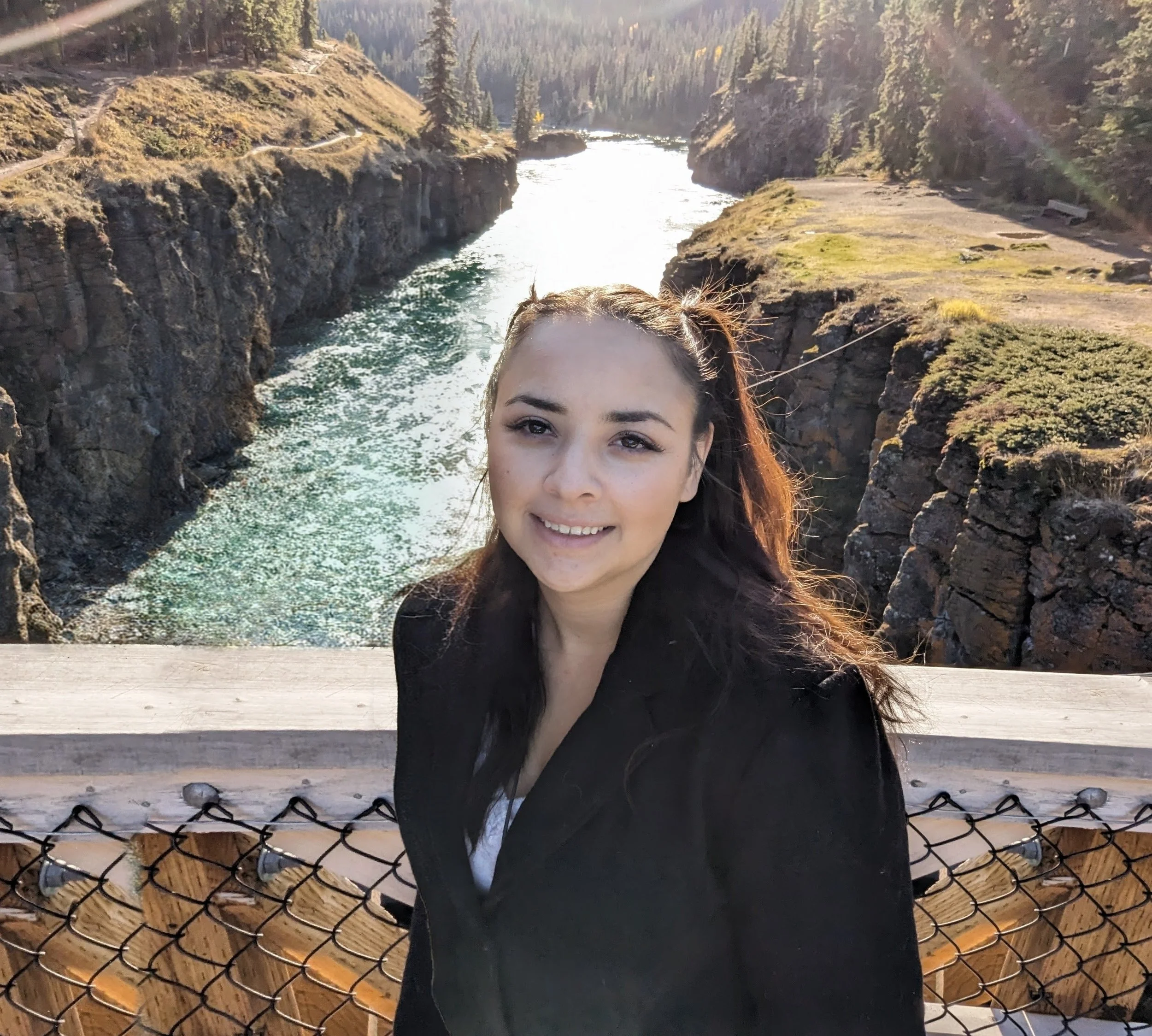 Woman with brown hair smiling in front of a river canyon with rocky cliffs and pine trees, wearing a black jacket and gray shoes on a wooden bridge with a chain-link fence.