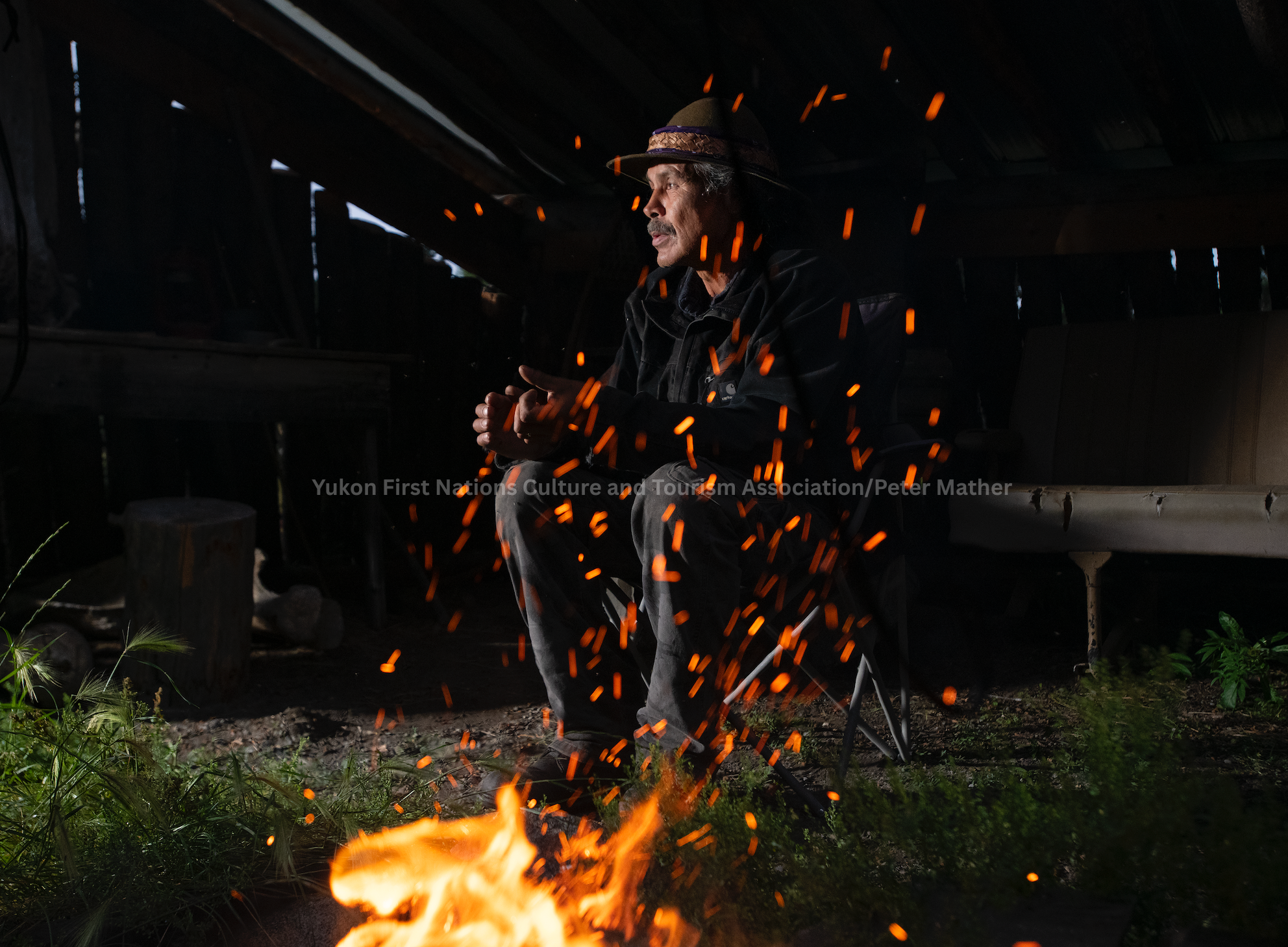 An older man with a mustache, wearing a wide-brimmed hat and a dark jacket, sitting by a campfire in a rustic wooden shelter at night, with glowing embers around him.