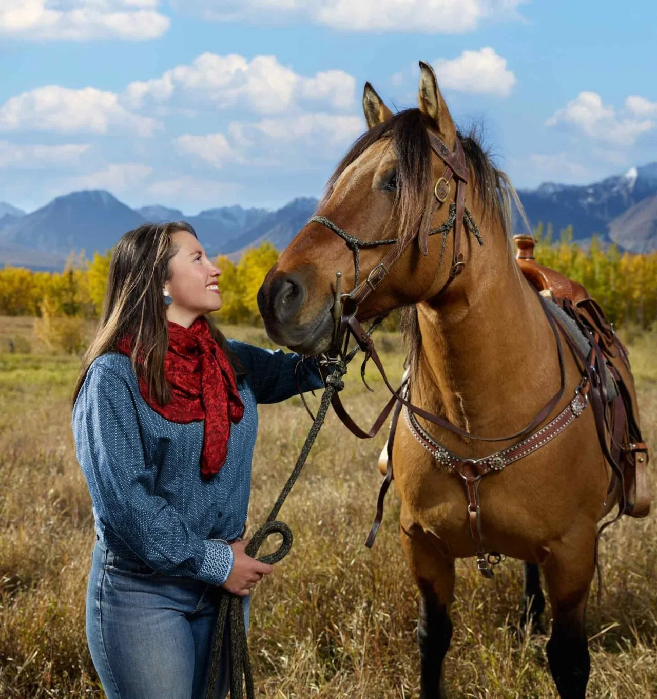 A woman in a blue shirt and red scarf standing in a field, smiling at a large brown horse with a saddle, mountains, and a partly cloudy sky in the background.