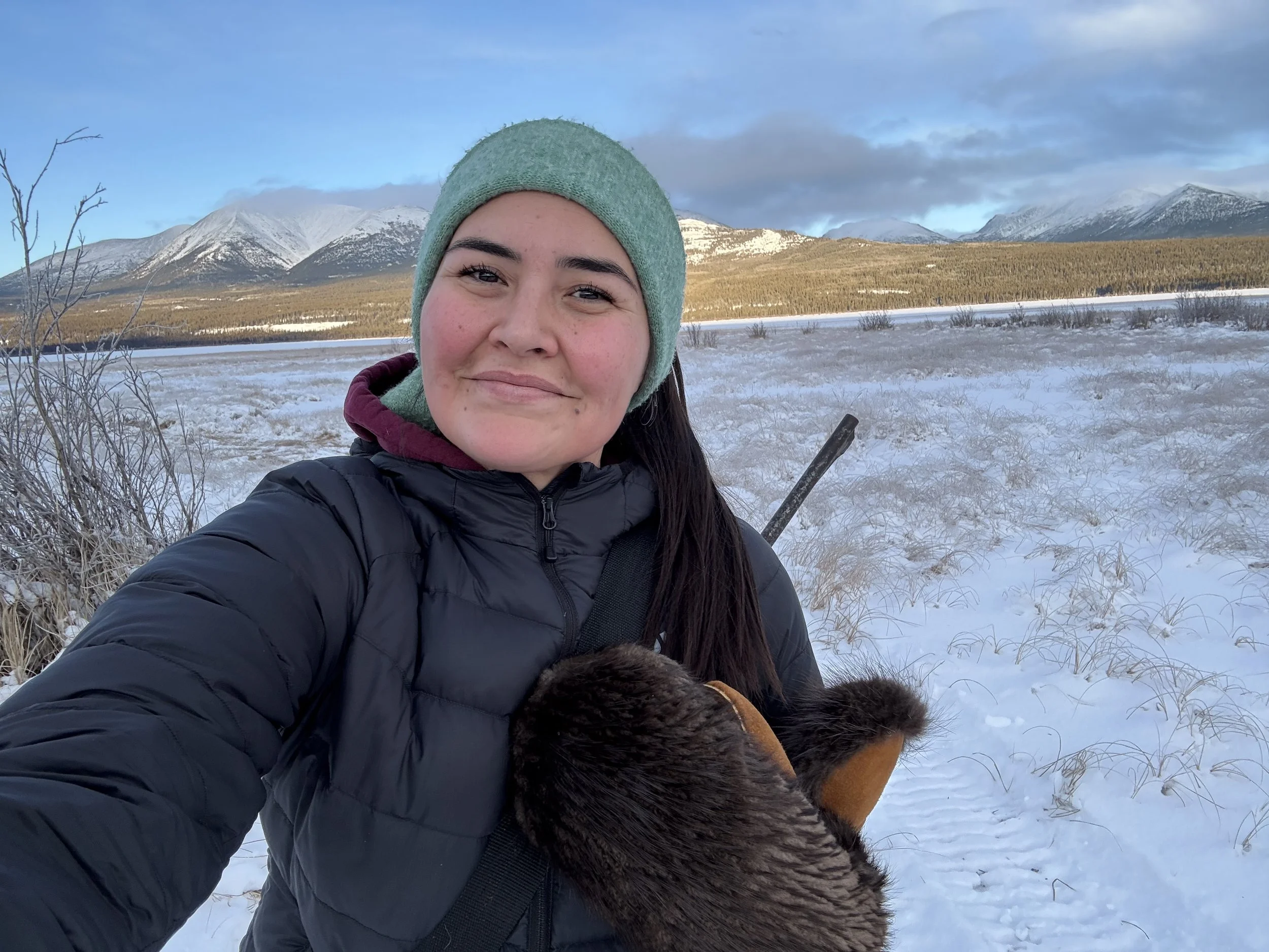 A woman wearing a green beanie and black jacket taking a selfie outdoors in a snowy landscape with mountains and blue sky in the background.