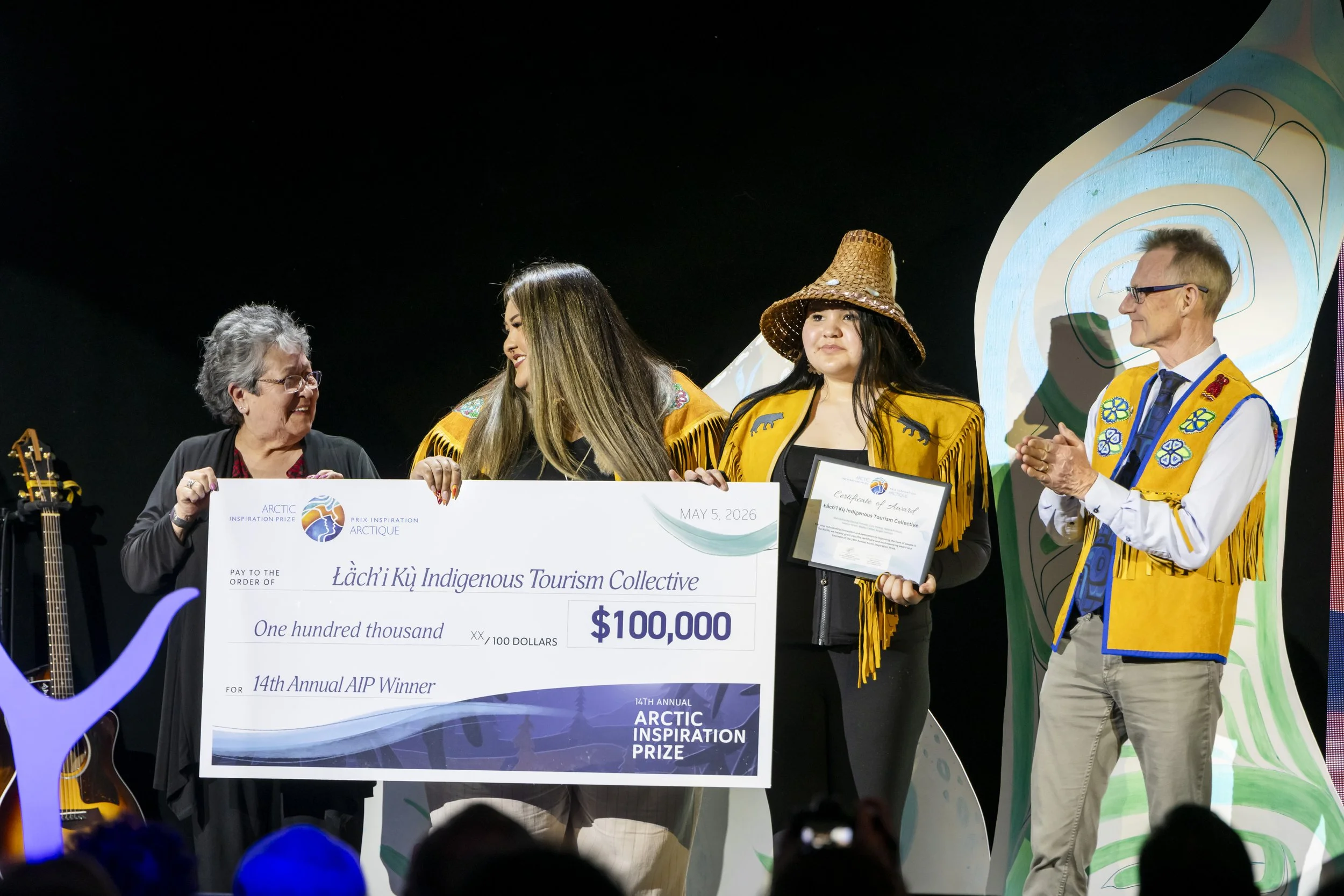 Group of award winners on stage receiving a large ceremonial check for $100,000 at the 14th Annual Arctic Inspiration Prize event, with participants wearing indigenous attire and holding certificates.