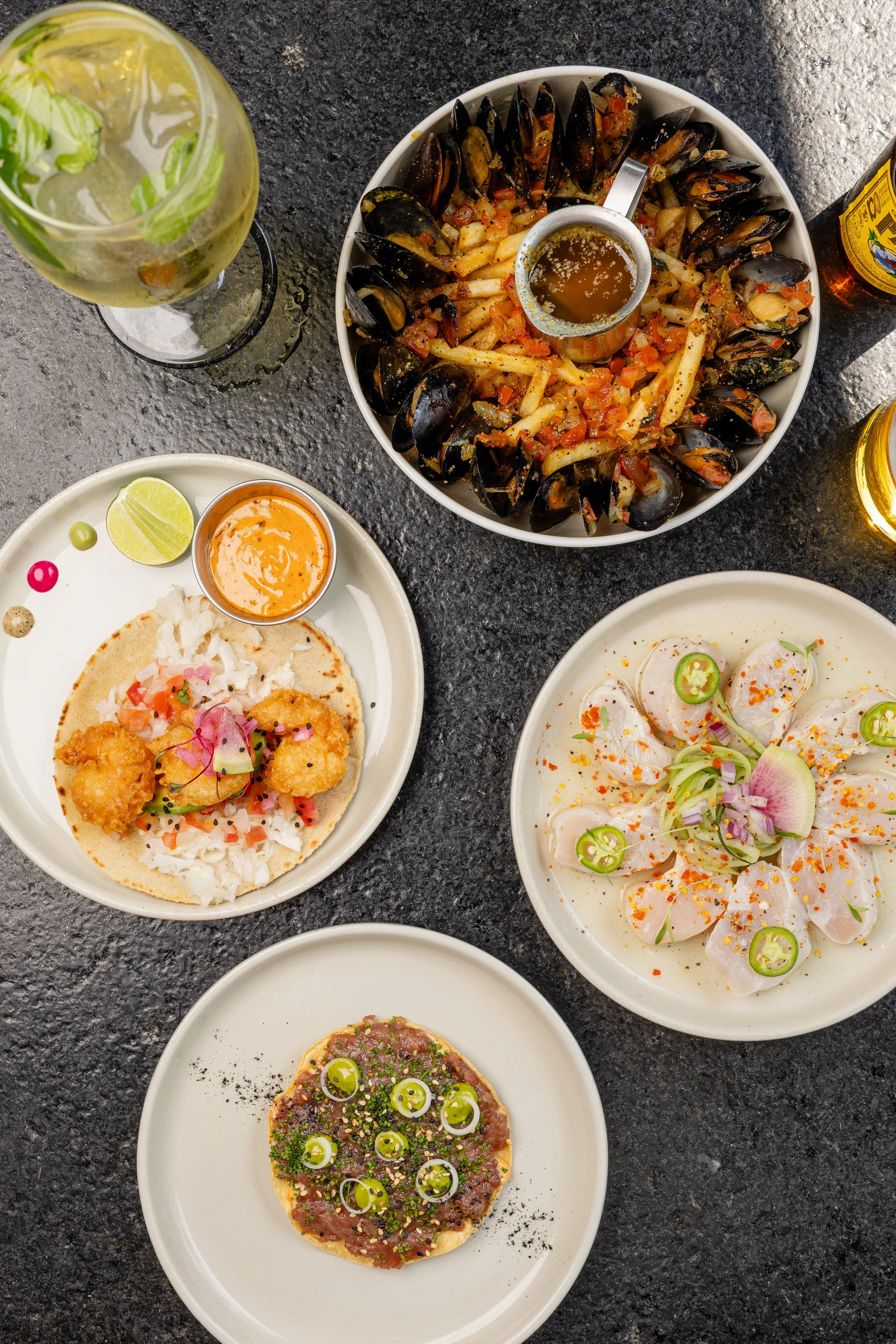A variety of Mexican dishes on white plates and bowls on a dark table, including ceviche, grilled steak with green onions, and tacos with rice, garnished with lime wedges, spicy sauces, and herbs, with glasses of beer and a glass of sparkling water.
