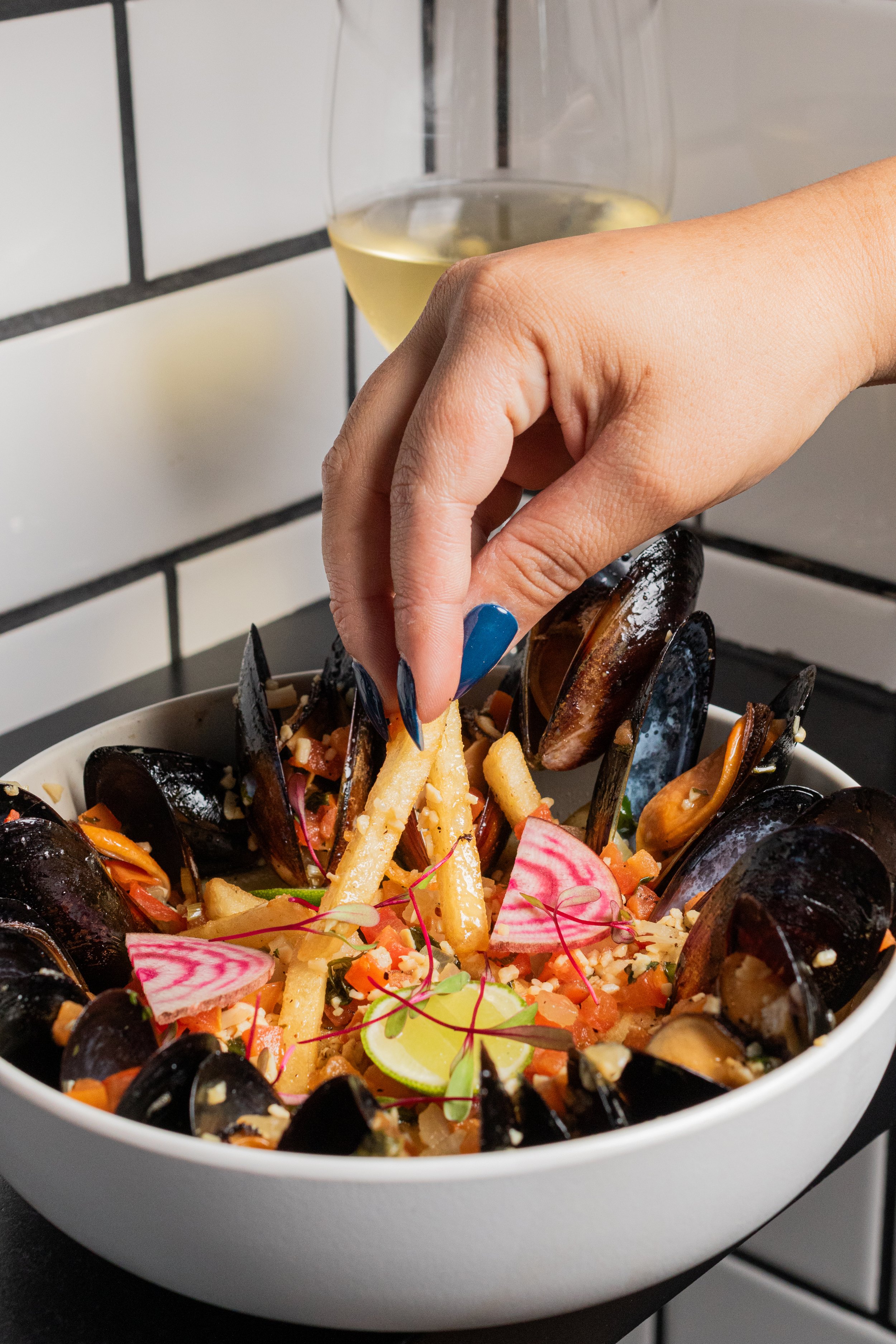 A bowl of seafood salad with mussels, chopped vegetables, and edible flowers, with a hand reaching to pick up a French fry, and a glass of white wine in the background.
