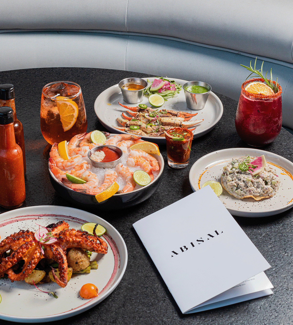 Assorted seafood dishes including shrimp with lemon wedges, octopus tentacles with garnishes, crab legs with sauces, and a seafood spread with herbs, along with cocktails garnished with citrus slices on a dark table.