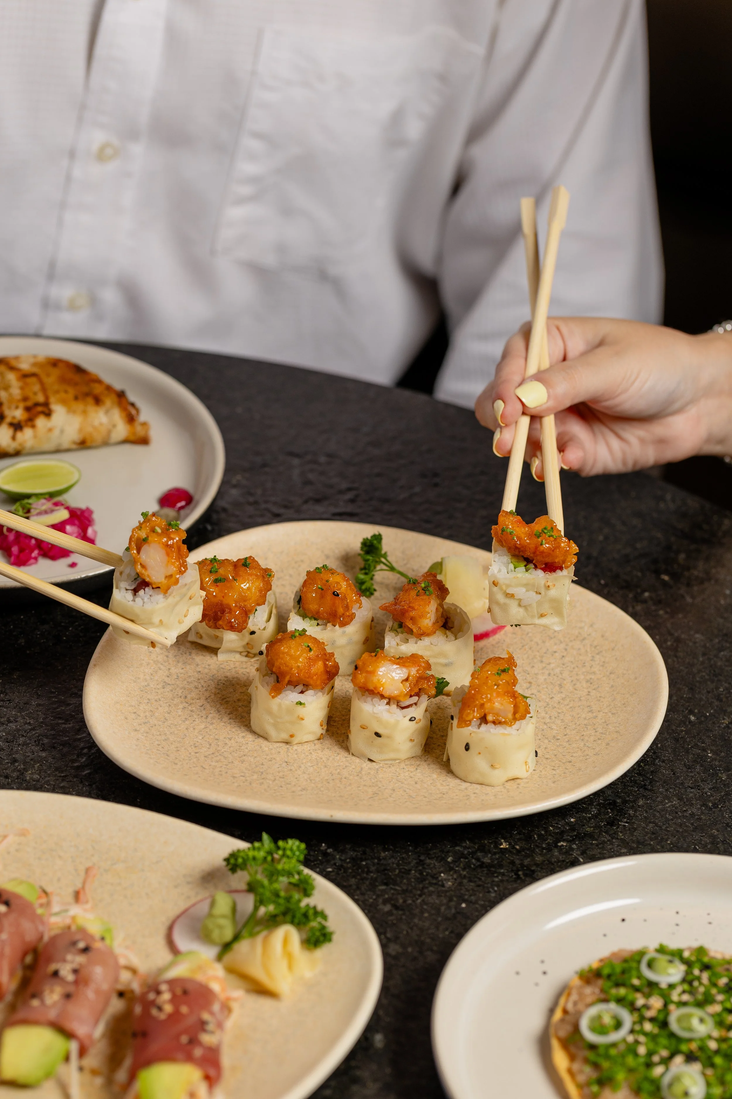 Close-up of a plate with sushi rolls topped with crispy shrimp, garnished with herbs, with chopsticks. Part of a pizza with cheese and toppings, and other dishes in the background.