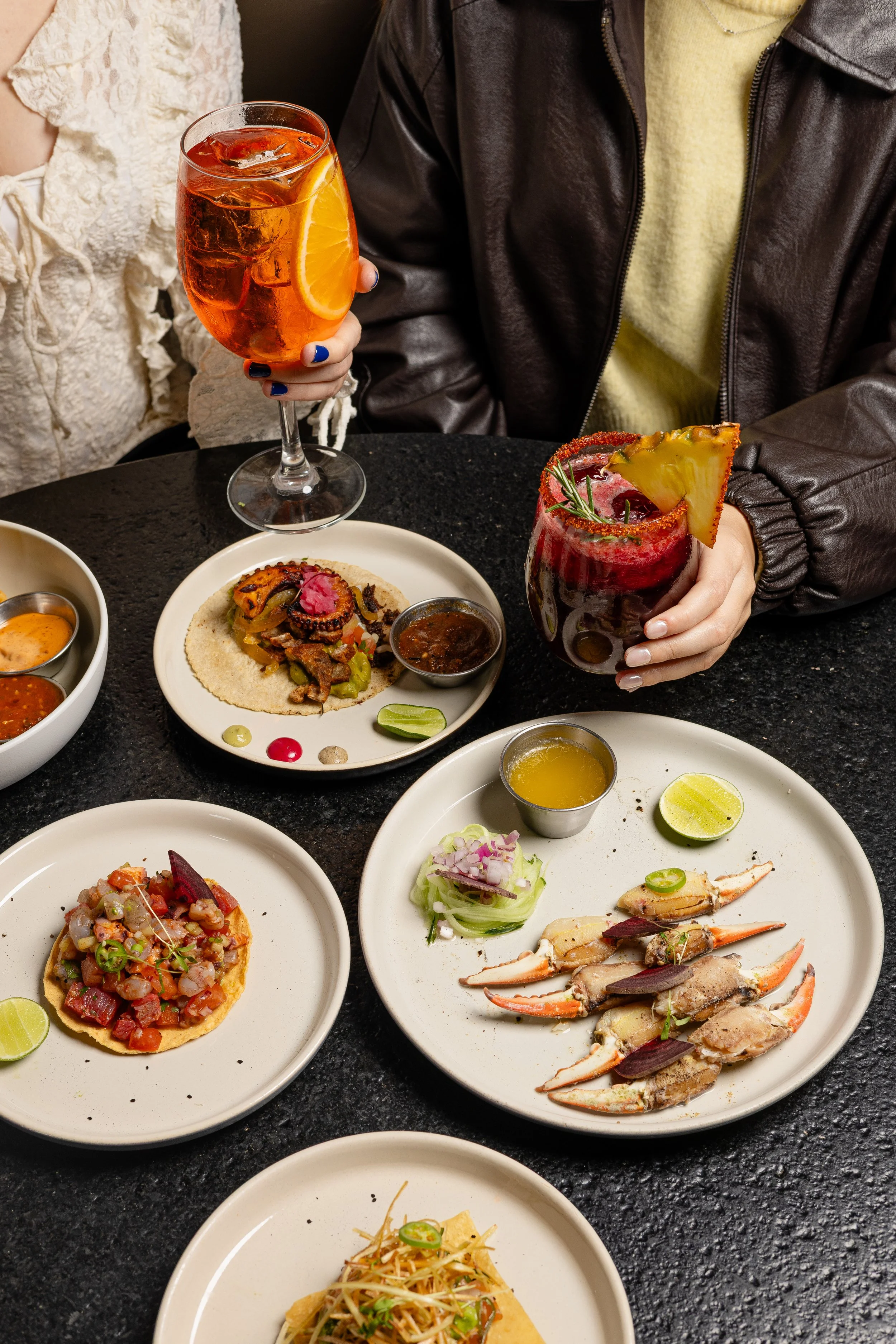 A table of various Mexican dishes and beverages, including a glass of orange-colored drink with ice and a slice of orange, a cocktail with pineapple and a salted rim, assorted tacos with toppings, garnished with lime slices and sauces.