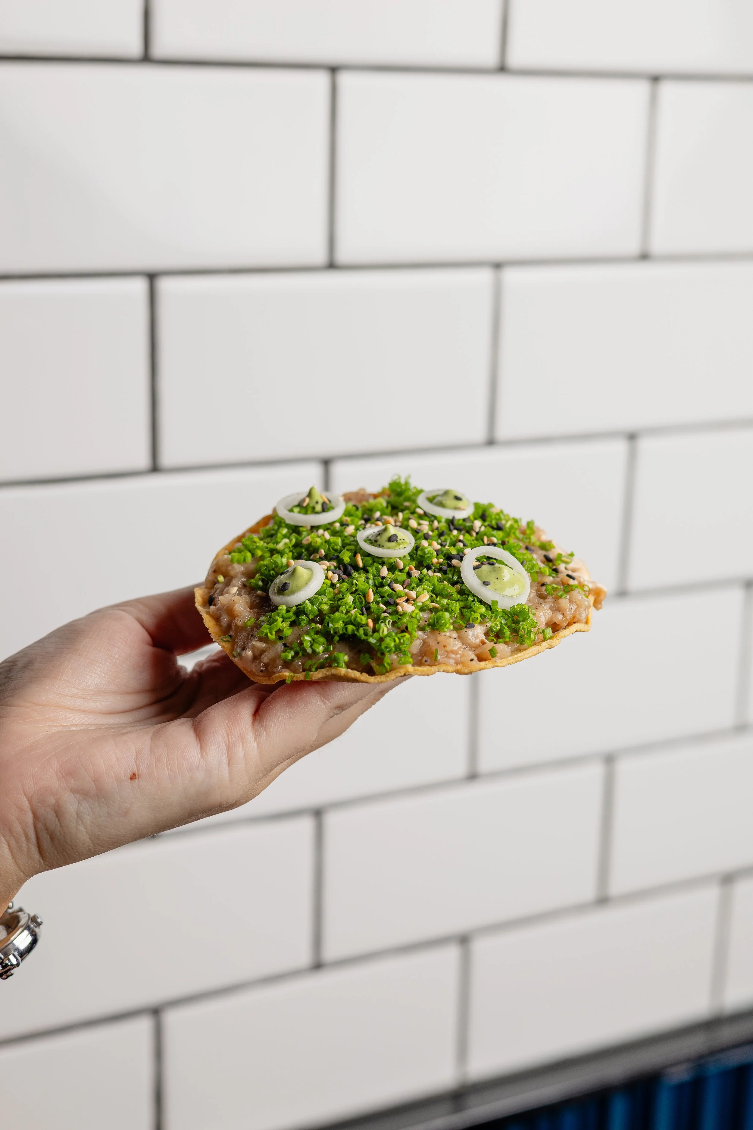 Close-up of a hand holding a slice of pizza topped with green herbs, onion slices, sesame seeds, and black seeds, against a white tiled wall background.
