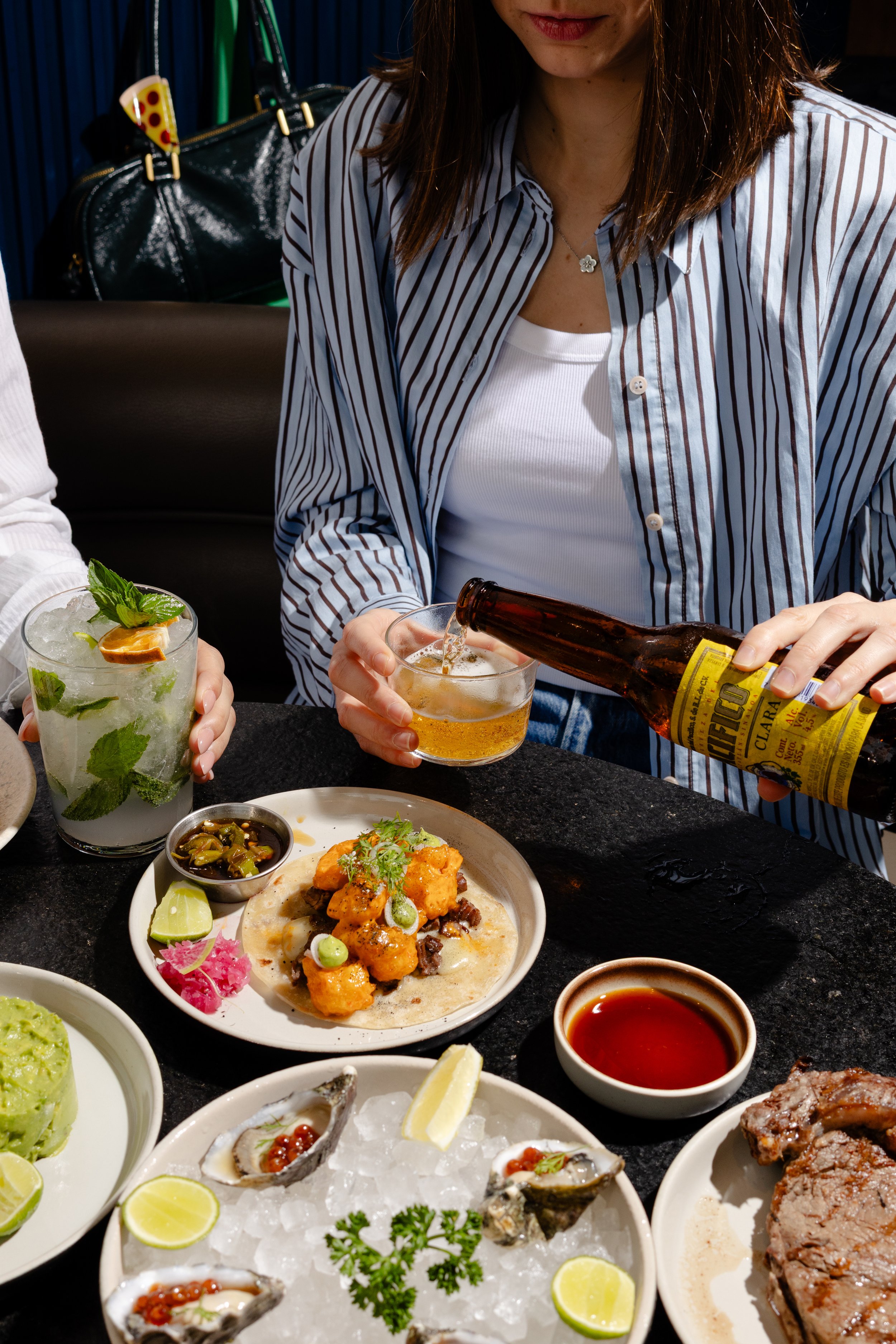 Close-up of a woman pouring a beer at a Mexican restaurant table with various Mexican dishes including oysters, guacamole, tacos, and chili peppers.