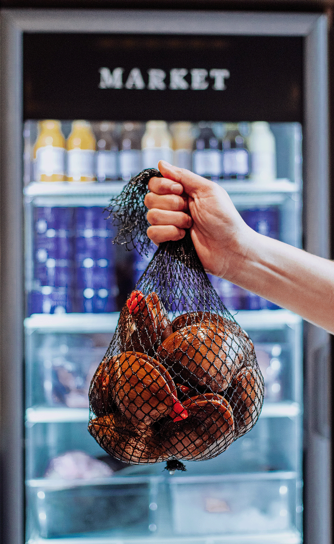 A hand holding a net bag of onions in front of a refrigerated display case at a market.
