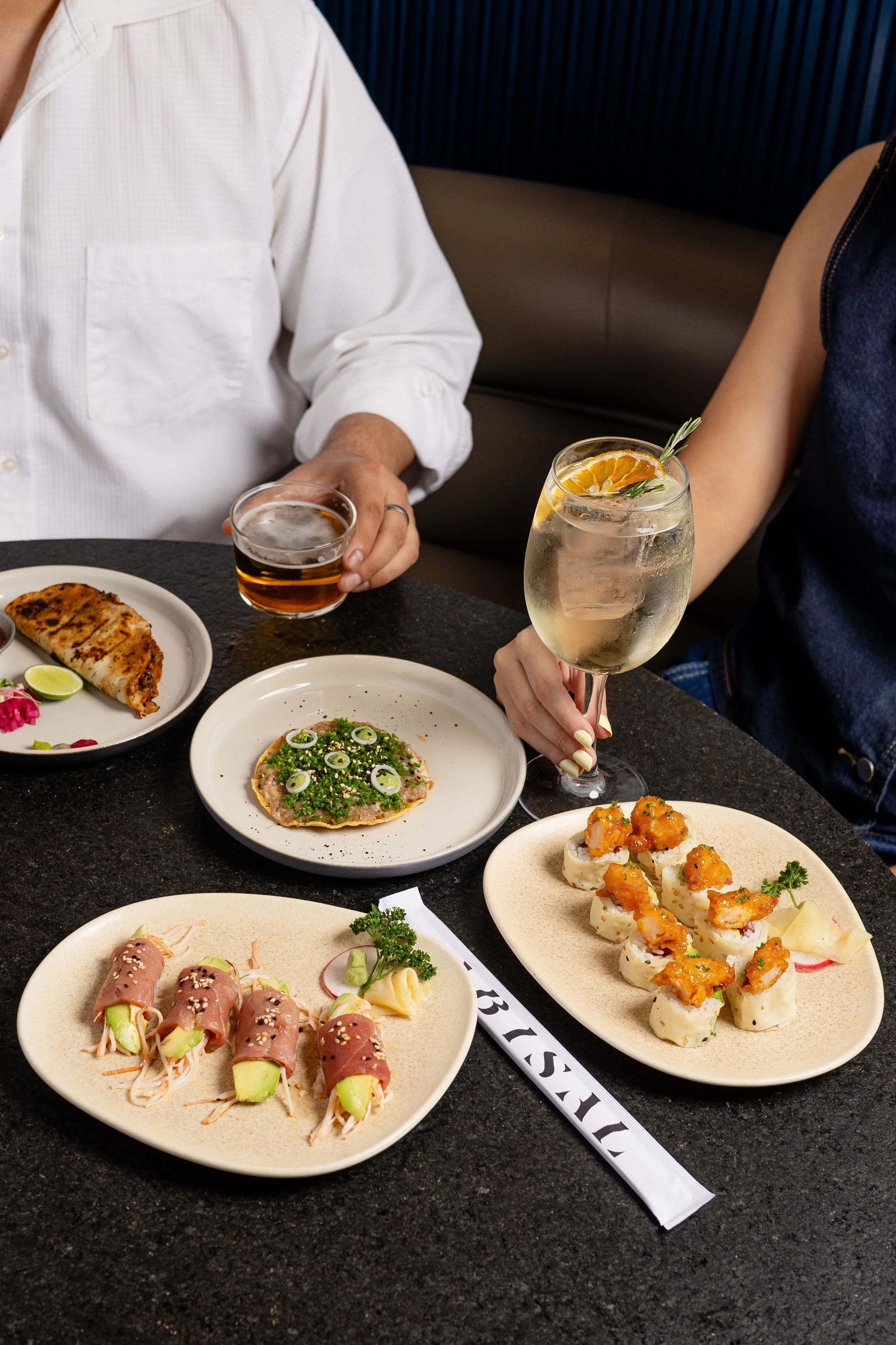 A group of people dining at a restaurant with a variety of sushi dishes and drinks on the table. The focus is on plates of sushi, and two people are holding drinks, one with a beer and the other with a cocktail garnished with orange slices and rosemary.